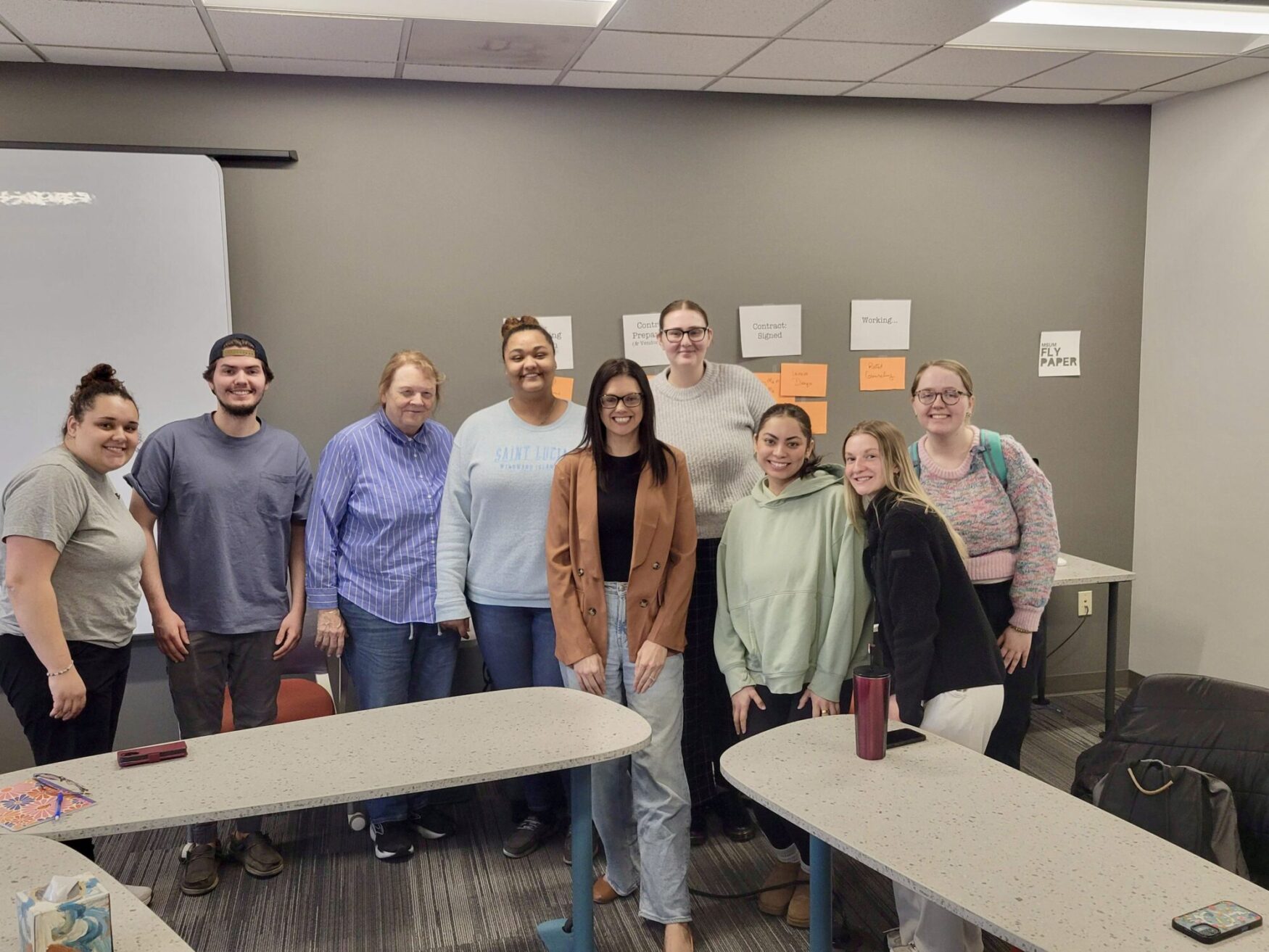 A group of nine people, consisting of men and women of varying ages and diverse ethnicities, pose together in a classroom. They are standing in front of a gray wall with several pieces of paper taped to it, containing handwritten labels like 'Contract Signed' and 'Working.' The room has whiteboard panels on one side and tables arranged in a U-shape. Some people are dressed casually in sweaters and jeans, while one person is wearing a blazer. They are all smiling and looking at the camera. There are notebooks, phones, and a red tumbler on the tables.
