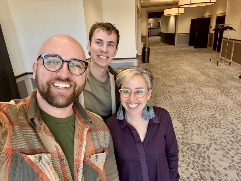 Three people stand together in a well-lit hallway, smiling at the camera. The person on the left is a bald man with glasses, a beard, and a warm smile, wearing a green shirt and an orange-and-brown plaid jacket. The person in the middle is a young man with short brown hair, wearing a green shirt and a light brown jacket, smiling brightly. The person on the right is a woman with short gray hair, wearing glasses, a purple button-up shirt, and large green tassel earrings. The background features a long hallway with neutral-colored walls, carpeted flooring, and overhead lighting.