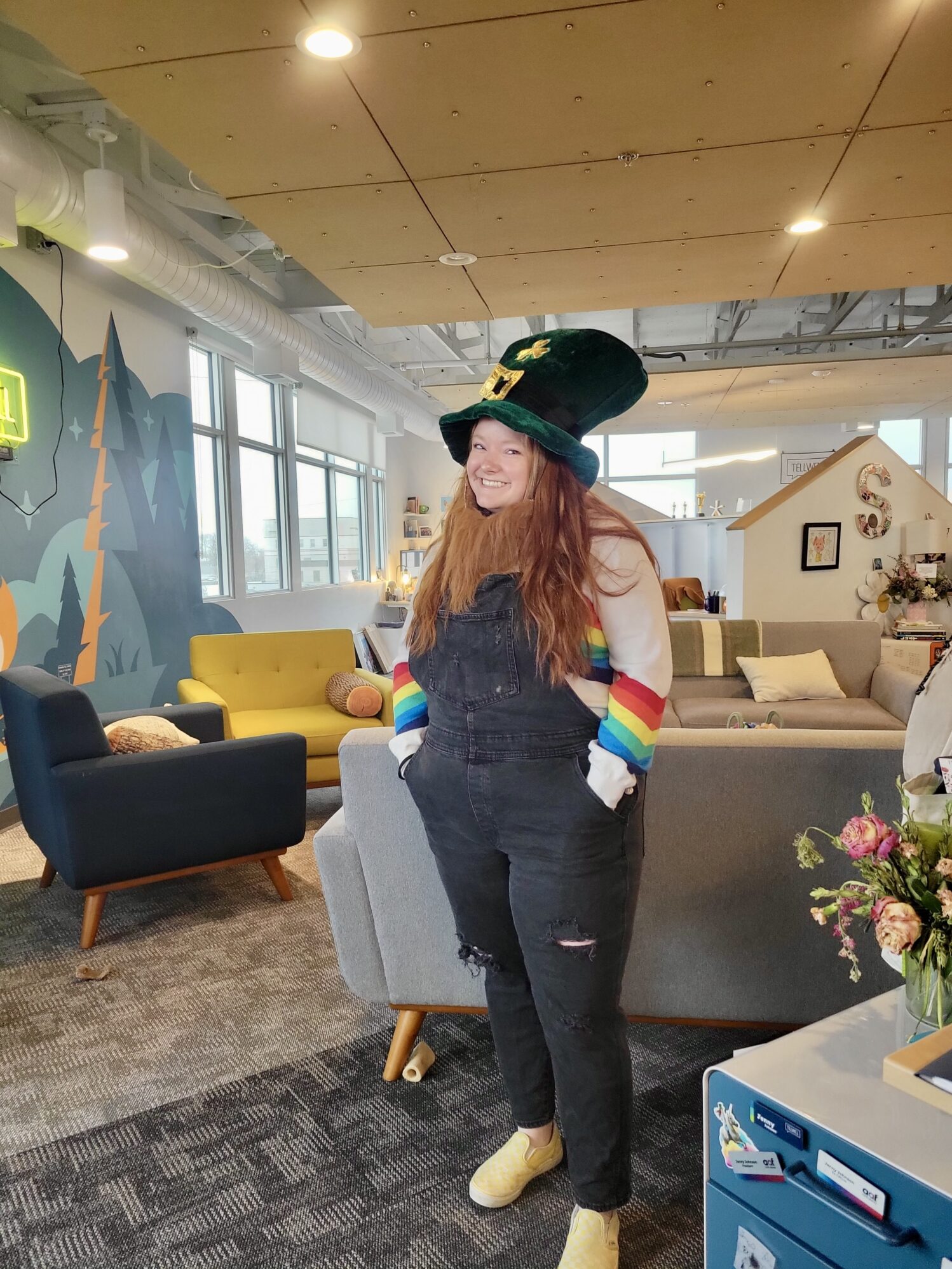A woman with long red hair stands in a brightly lit, modern office space, smiling at the camera. She wears a large green leprechaun hat with a gold buckle and shamrock, along with black overalls over a white long-sleeve shirt that has rainbow-striped sleeves. Her hands are tucked into her pockets, and she wears yellow checkered slip-on shoes. The office features colorful furniture, a mural with trees and mountains, and a cozy, creative atmosphere with various decorations and plants.