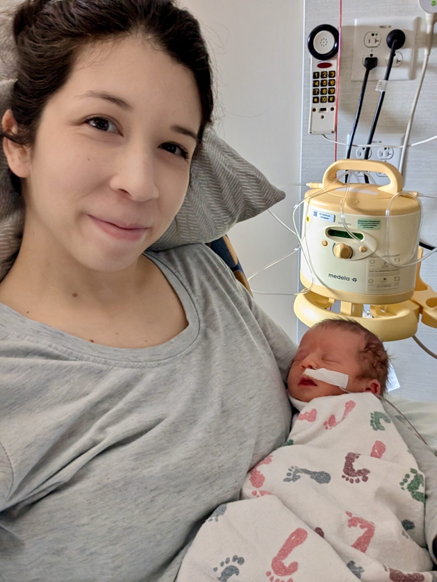 A woman with dark hair pulled back is smiling softly while holding a newborn baby in her arms. The baby is wrapped in a white hospital blanket with colorful footprints and has a small nasal tube for oxygen support. They are in a hospital room, with medical equipment including a breast pump and wall-mounted devices visible in the background. The woman is wearing a light gray t-shirt and is resting against a pillow, looking at the camera.