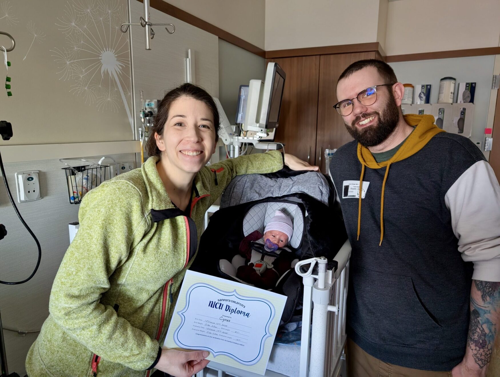 A woman and a man are smiling and posing with their newborn baby in a hospital room. The baby is snug in a car seat, wearing a striped hat and using a pacifier. The woman is holding a "NICU Diploma," indicating the baby has graduated from the Neonatal Intensive Care Unit. She is wearing a green fleece jacket, and the man, who has glasses and a beard, is dressed in a gray and yellow hoodie. Medical equipment is visible in the background, including monitors and IV stands, along with decorative dandelion wall art.