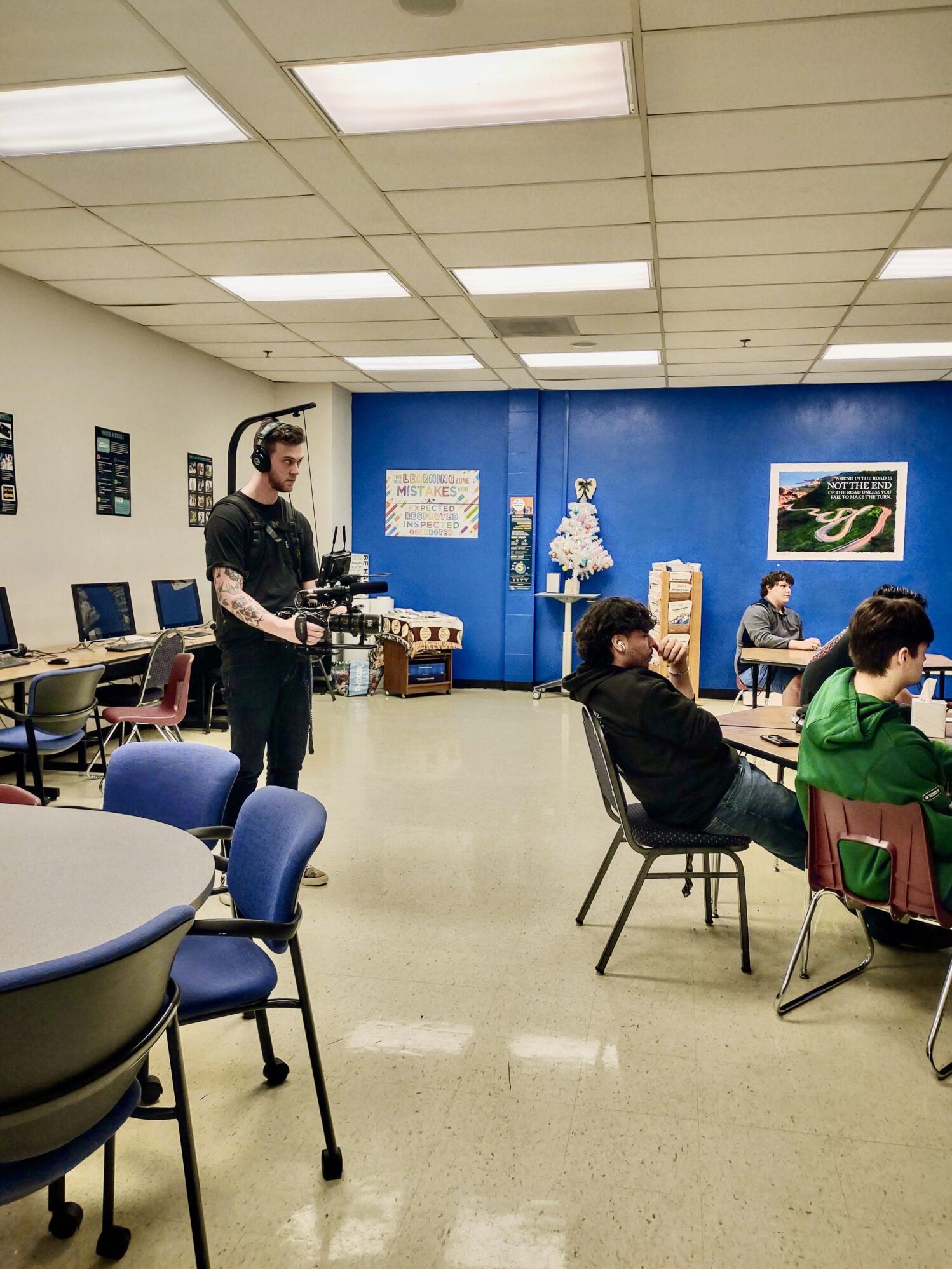 A videographer wearing headphones and holding a professional camera rig films inside a classroom with blue and white walls. Several students are seated at tables, engaged in conversation or working on tasks. Computers line one side of the room, and motivational posters and decorations adorn the walls. The setting appears to be an educational or community space.
