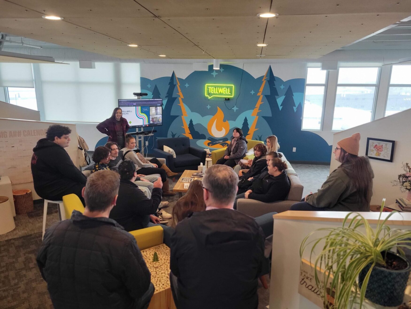 This image captures a group of people gathered in a modern, casual office space for a discussion or presentation. The room features cozy seating, including couches and stools arranged in a semi-circle. A woman at the front stands next to a screen displaying a presentation. The background includes a vibrant mural with a campfire, trees, and a neon sign reading "Tellwell," adding a creative and inviting atmosphere. The space is well-lit with large windows allowing natural light to stream in, and there are personal touches like plants and artwork on the walls.