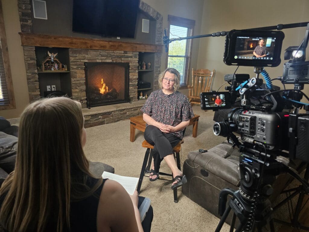 A woman sits on a stool in a living room being filmed for an interview, with a fireplace in the background and camera equipment in the foreground.