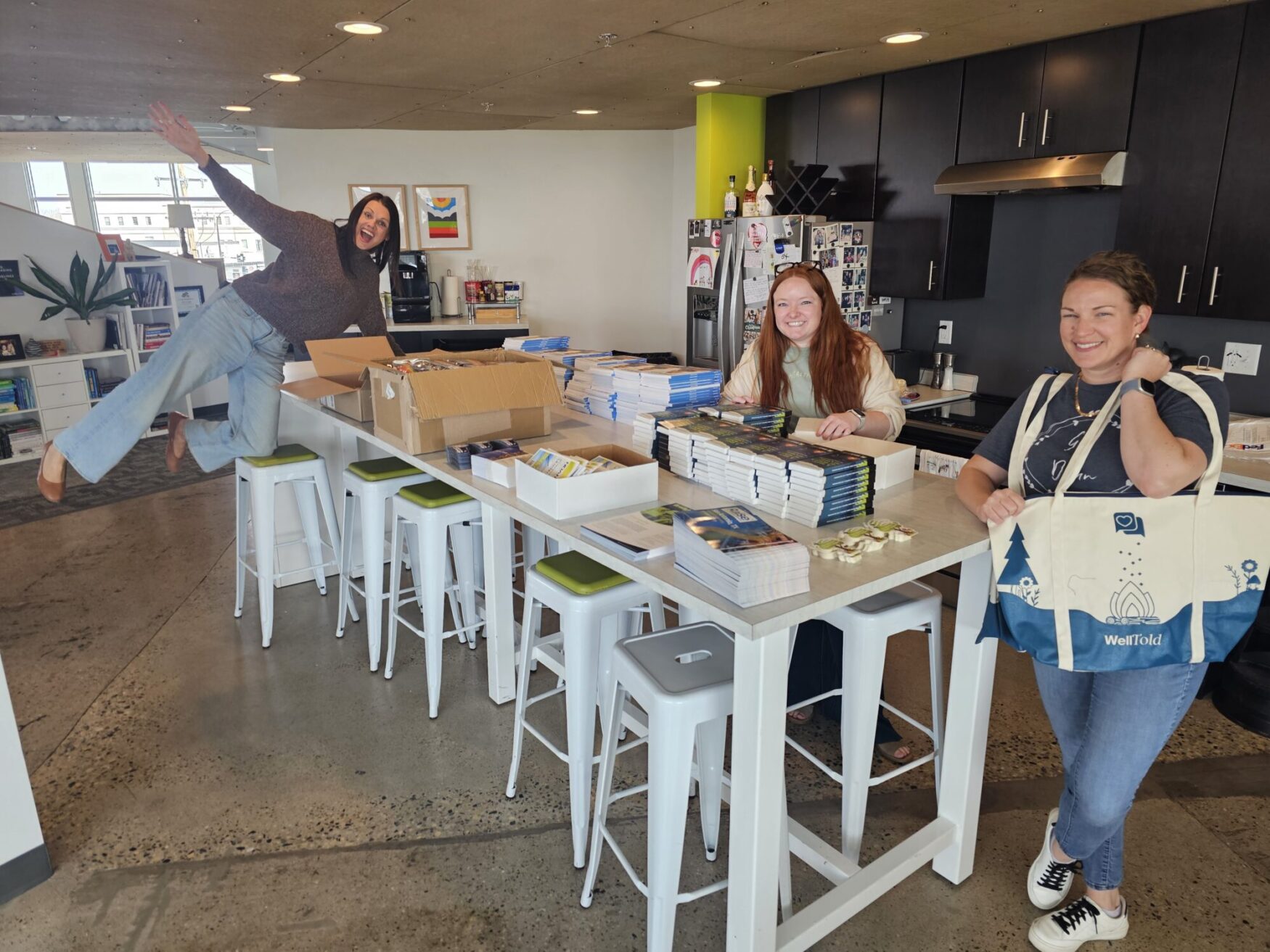 Three women smile as they prepare event materials at a kitchen island; one woman playfully poses mid-leap while another holds a WellTold tote bag filled with supplies.