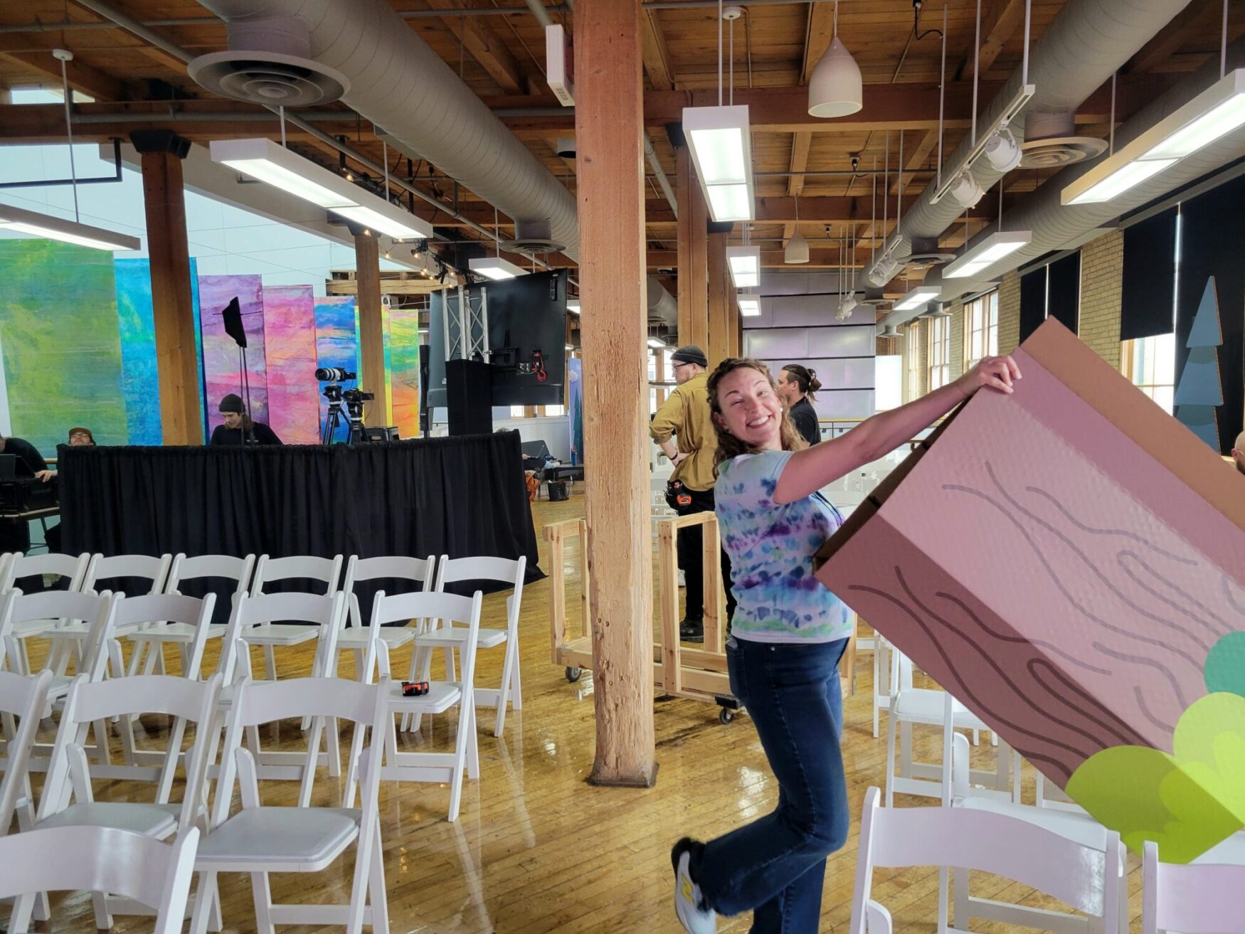 A woman in a tie-dye shirt playfully poses while carrying a large cardboard cutout resembling a tree trunk, surrounded by white chairs and a colorful event setup in progress.