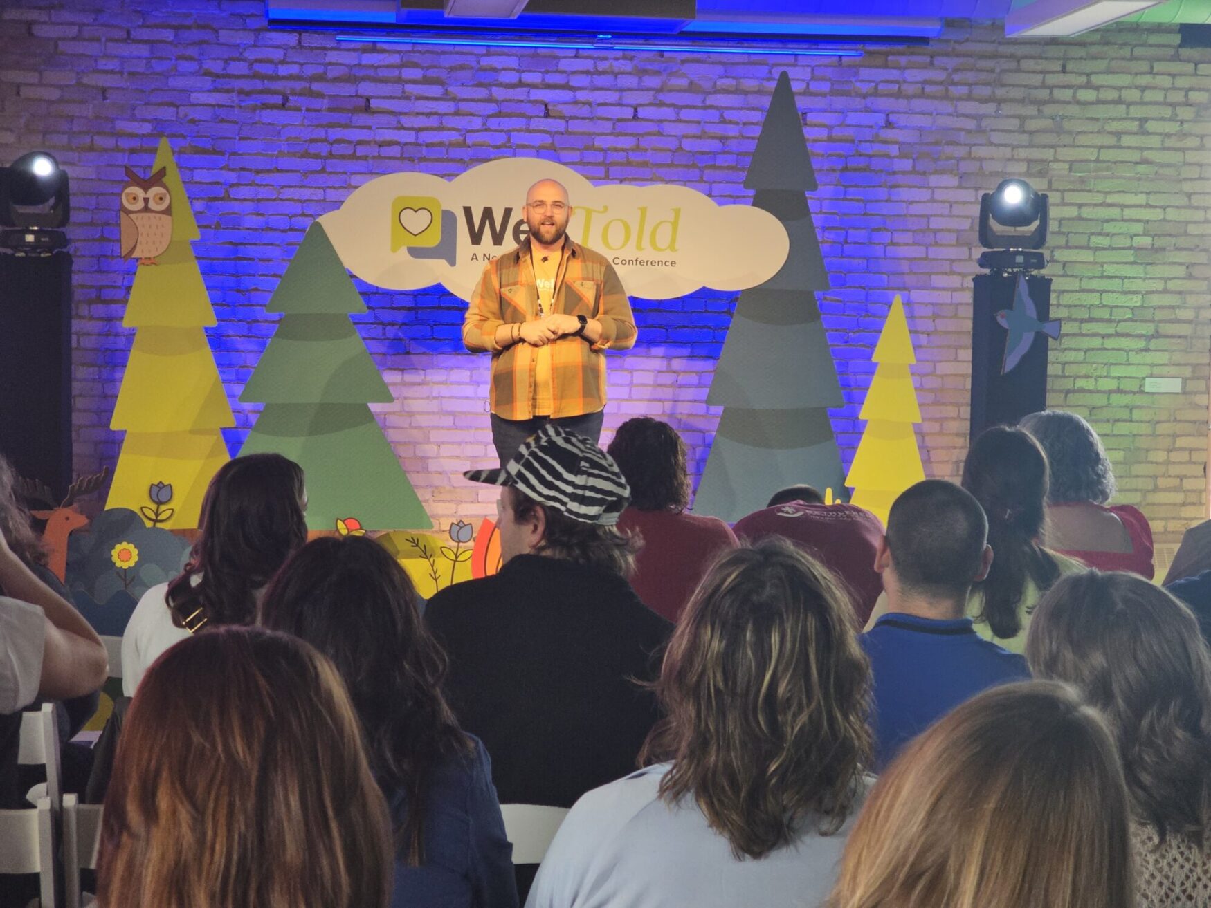 A speaker stands on a colorful stage at the WellTold conference, framed by whimsical tree and animal cutouts, addressing an attentive seated audience.
