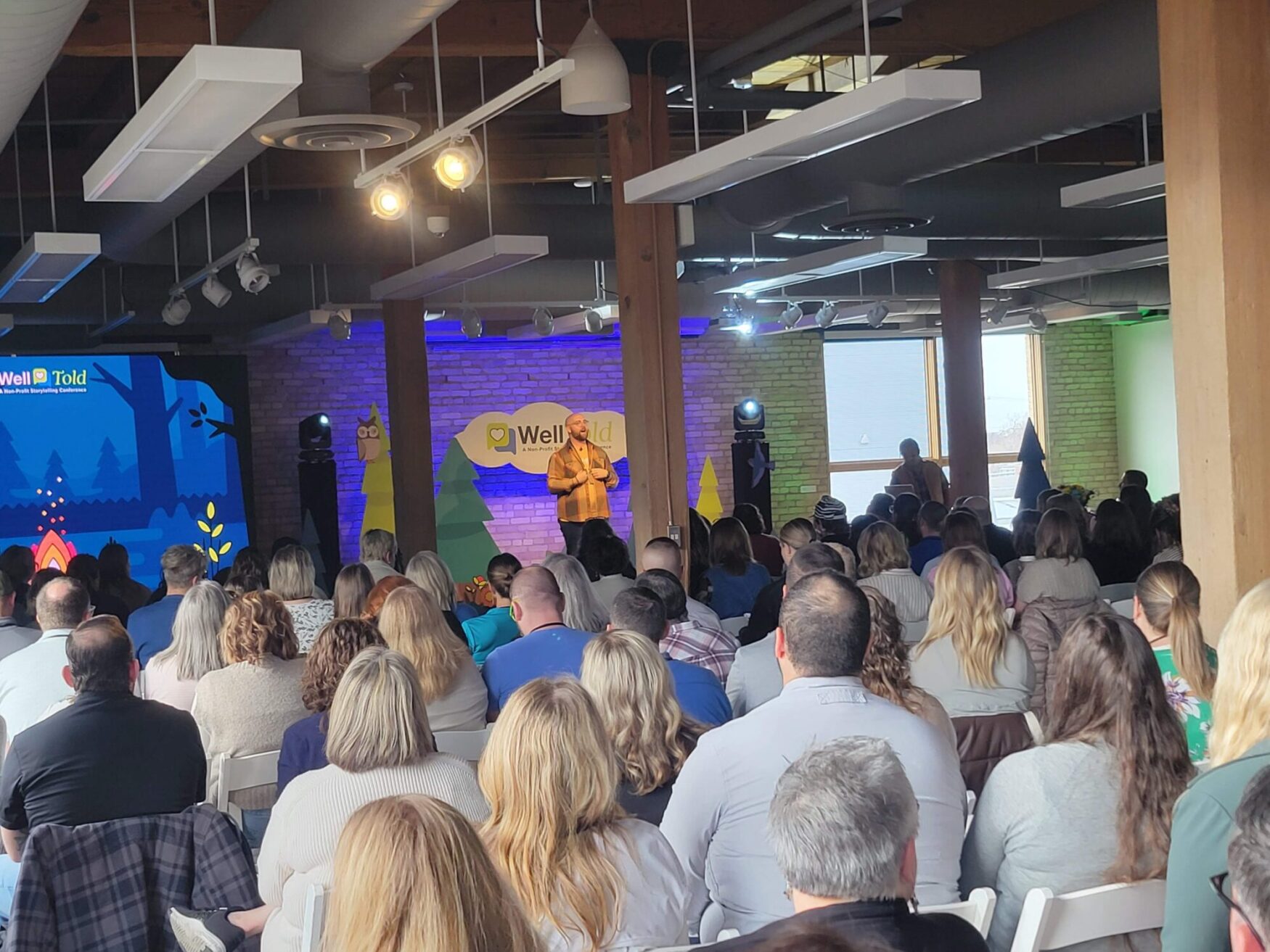 A speaker presents on stage at the WellTold storytelling conference, with a large audience seated and the colorful conference branding and woodland-themed decorations in the background.