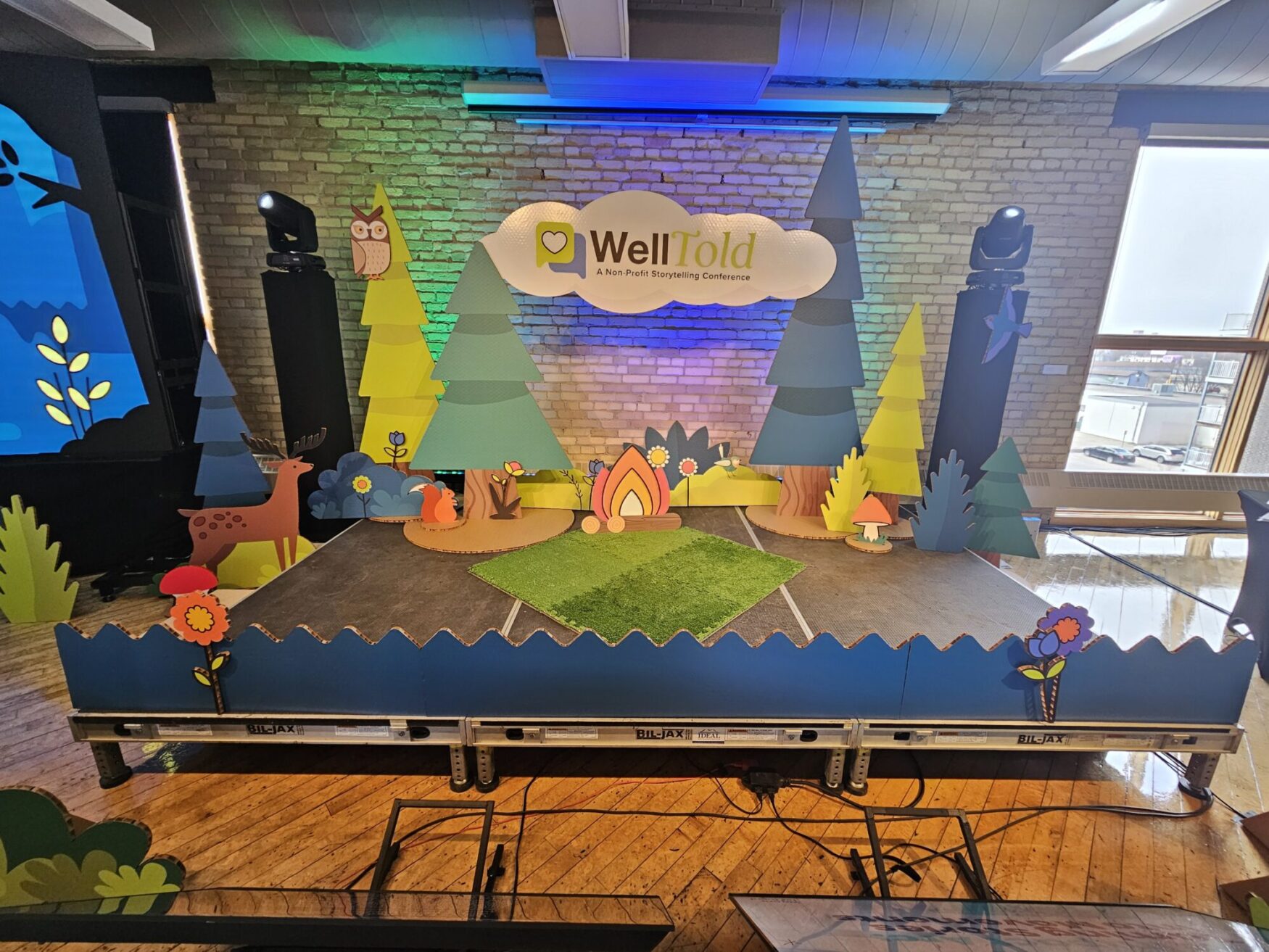 A whimsical stage set for the WellTold conference, decorated with colorful cardboard cutouts of trees, animals, and a central campfire theme.