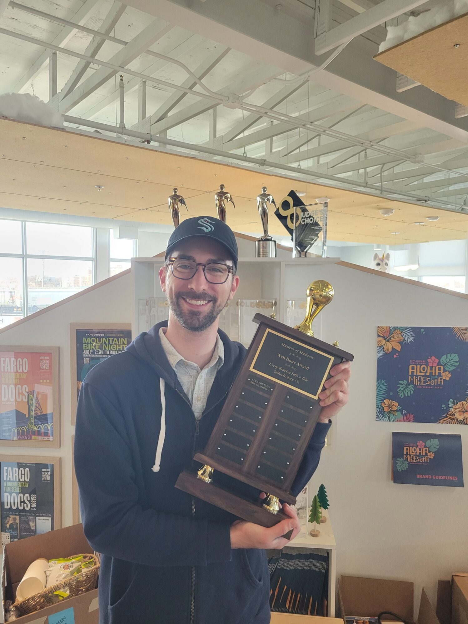 A smiling man in a cap and glasses proudly holds a large trophy labeled 'Well Done Award' inside a creative office space, with award trophies and colorful posters in the background.