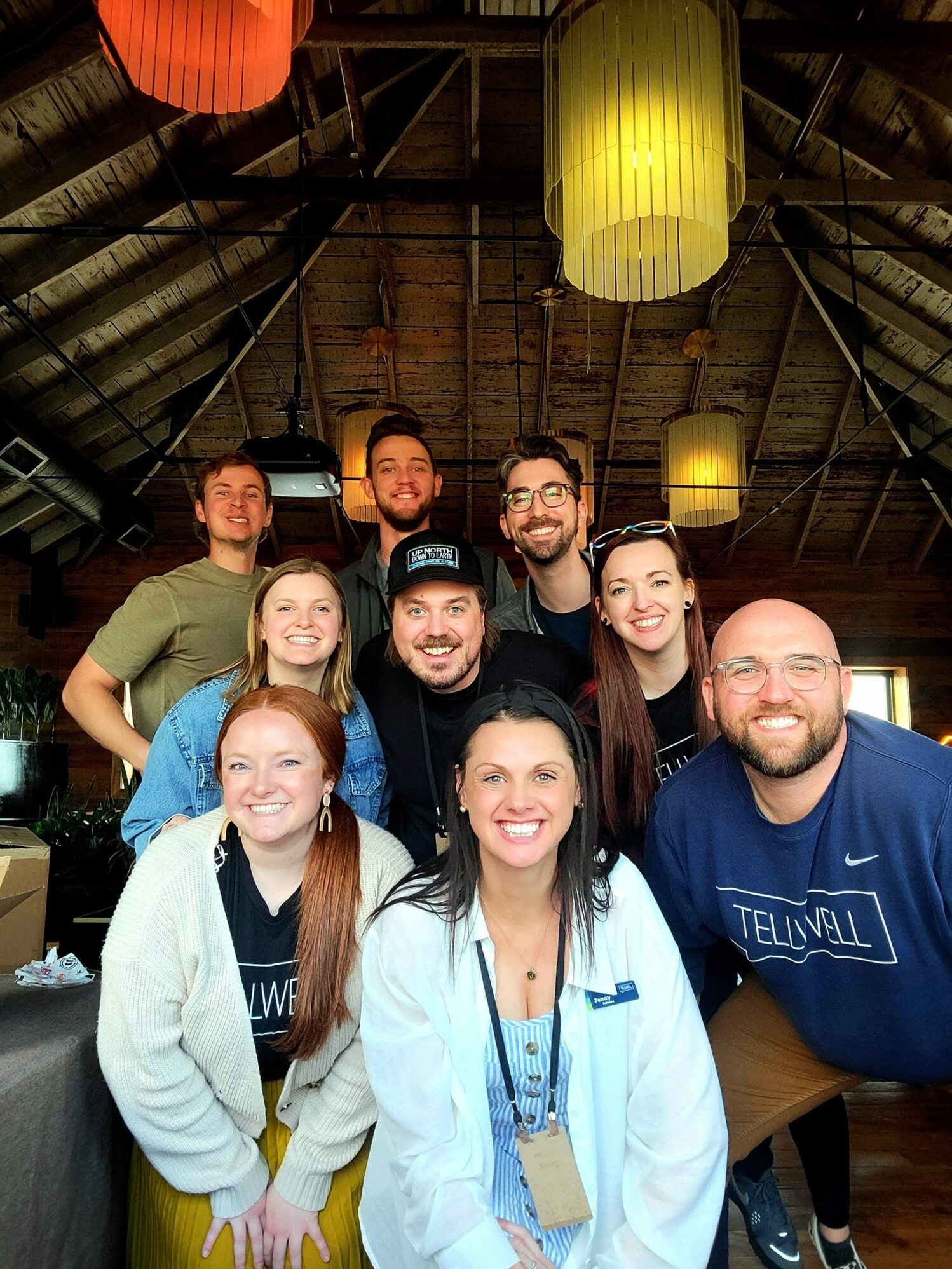 A cheerful group photo of the Tellwell team posing together under warm hanging lights in a cozy, wooden-ceilinged venue, all smiling and dressed in branded gear.