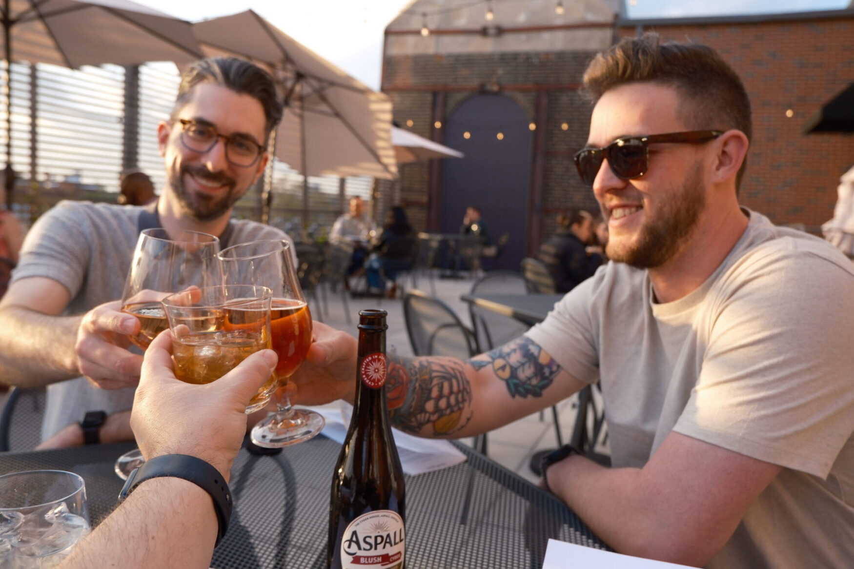Three people sitting at an outdoor table clink glasses while smiling. There are drinks and a bottle labeled "Aspall" on the table. Other people and umbrellas are in the background.