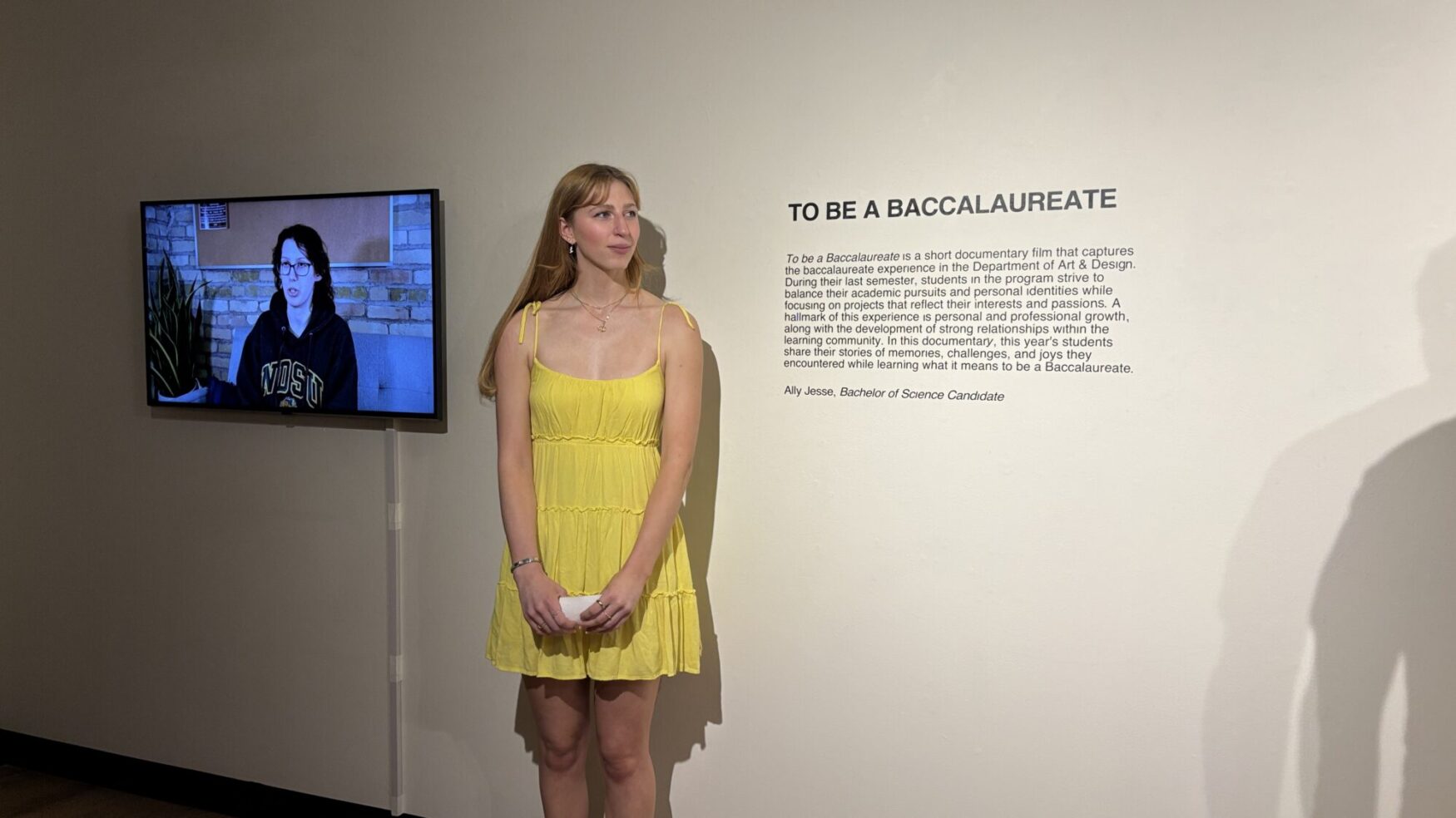 A girl with red hair and a yellow dress stands in front of a TV screen holding note cards, appearing to be presenting to a group. The wall reads “To Be A Baccalaureate” with additional text underneath. This appears to be in an art gallery.