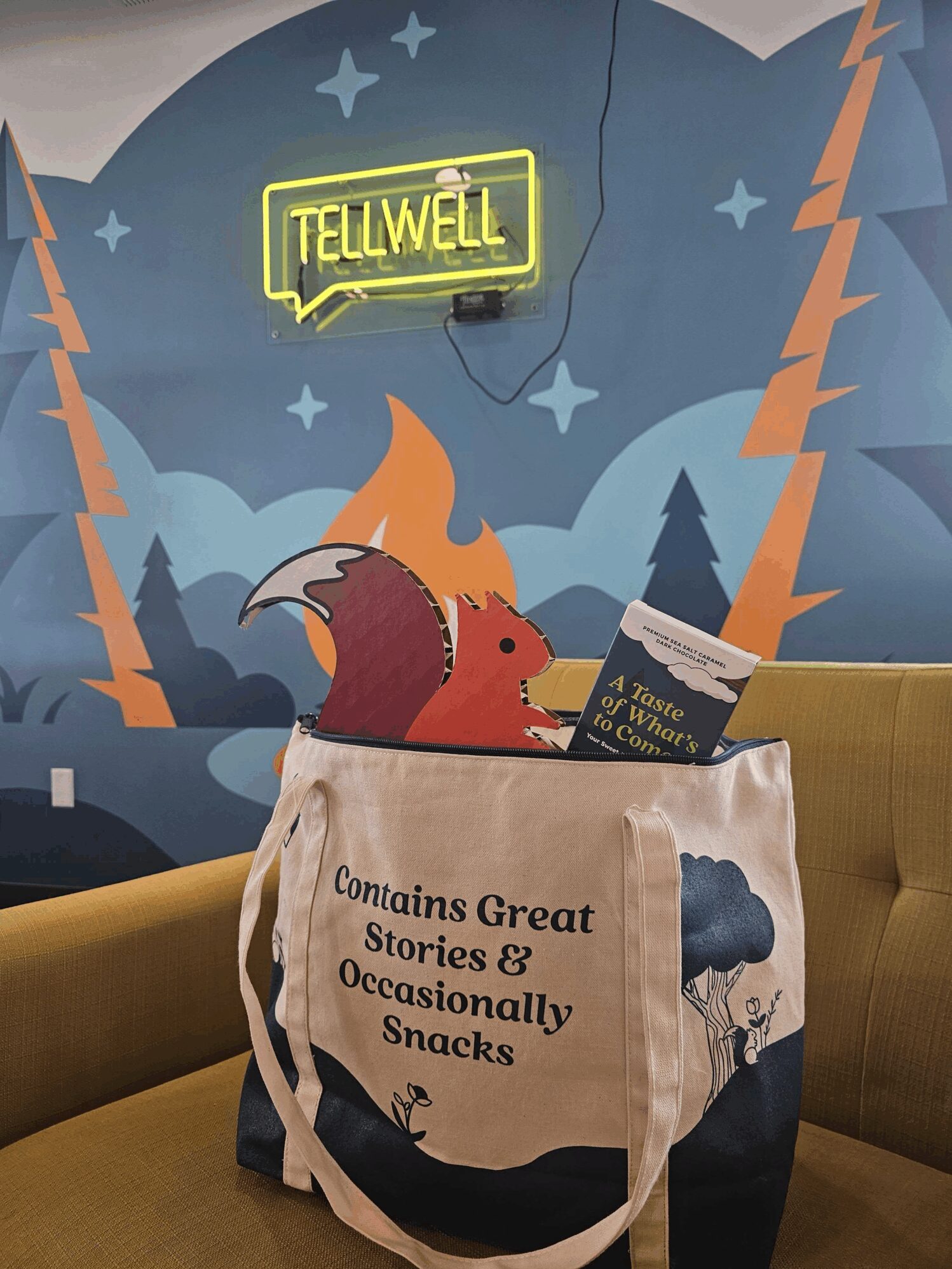 A WellTold-branded binder with a storytelling quote and a matching canvas tote bag sit on a table, with blurred attendees mingling in the background of a warmly lit venue.