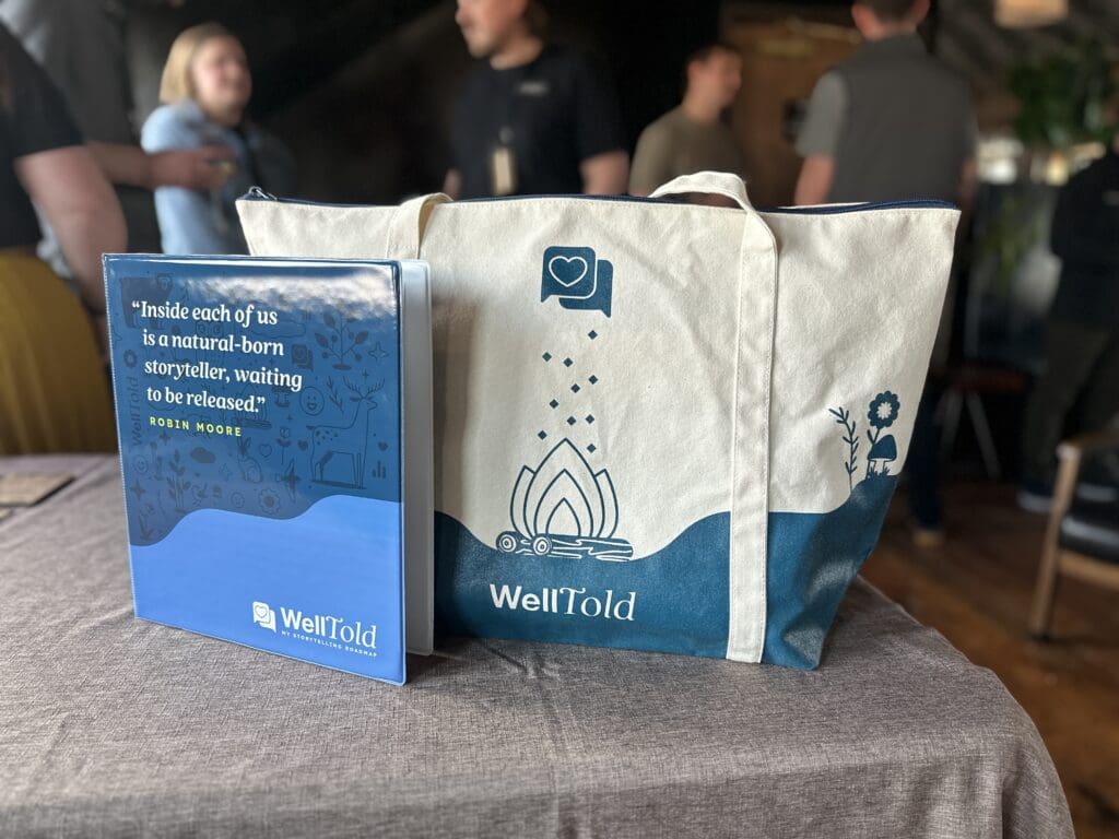 A WellTold-branded binder with a storytelling quote and a matching canvas tote bag sit on a table, with blurred attendees mingling in the background of a warmly lit venue.
