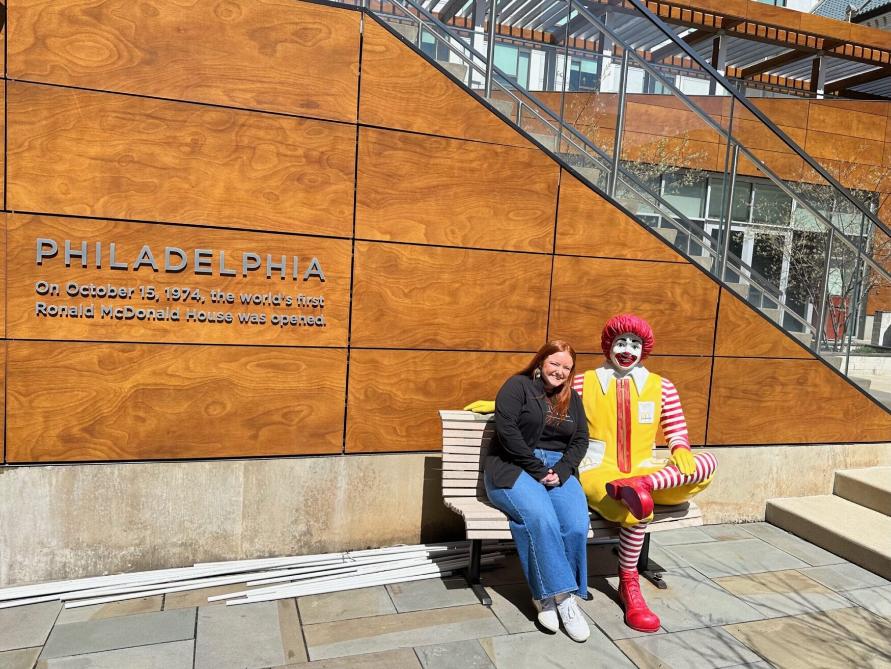 A smiling woman sits on a bench next to a statue of Ronald McDonald, leaning slightly toward it. Behind them, a large wooden wall features an inscription about the world's first Ronald McDonald House, which opened in Philadelphia on October 15, 1974. A modern staircase and glass railing add depth to the background.