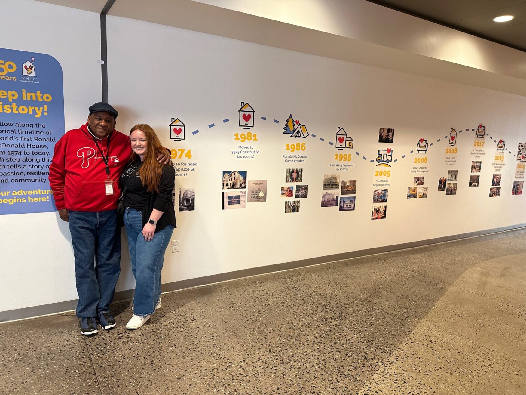 A woman and a man stand together, smiling in front of a historical timeline display at the Ronald McDonald House in Philadelphia. The man wears a red Philadelphia Phillies hoodie and a lanyard, while the woman wears a black top and jeans. The timeline on the wall highlights key milestones in the organization's history, beginning in 1974 and continuing through the years.