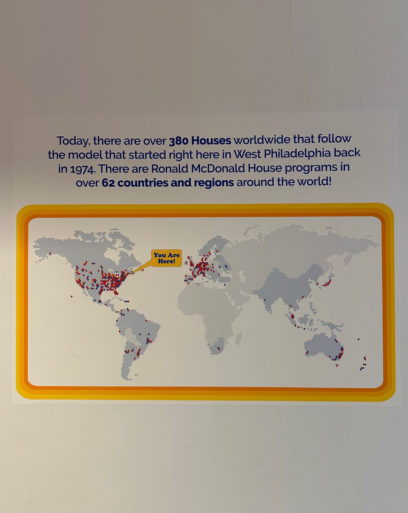 This image features an informational sign about Ronald McDonald House programs worldwide. The sign states that there are over 380 Houses globally, following a model that began in West Philadelphia in 1974. It highlights that these programs exist in 62 countries and regions. Below the text, there is a world map with red dots marking the locations of Ronald McDonald Houses. A yellow label with "You Are Here!" points to a specific location in the United States. The sign is bordered with a yellow and orange gradient frame.