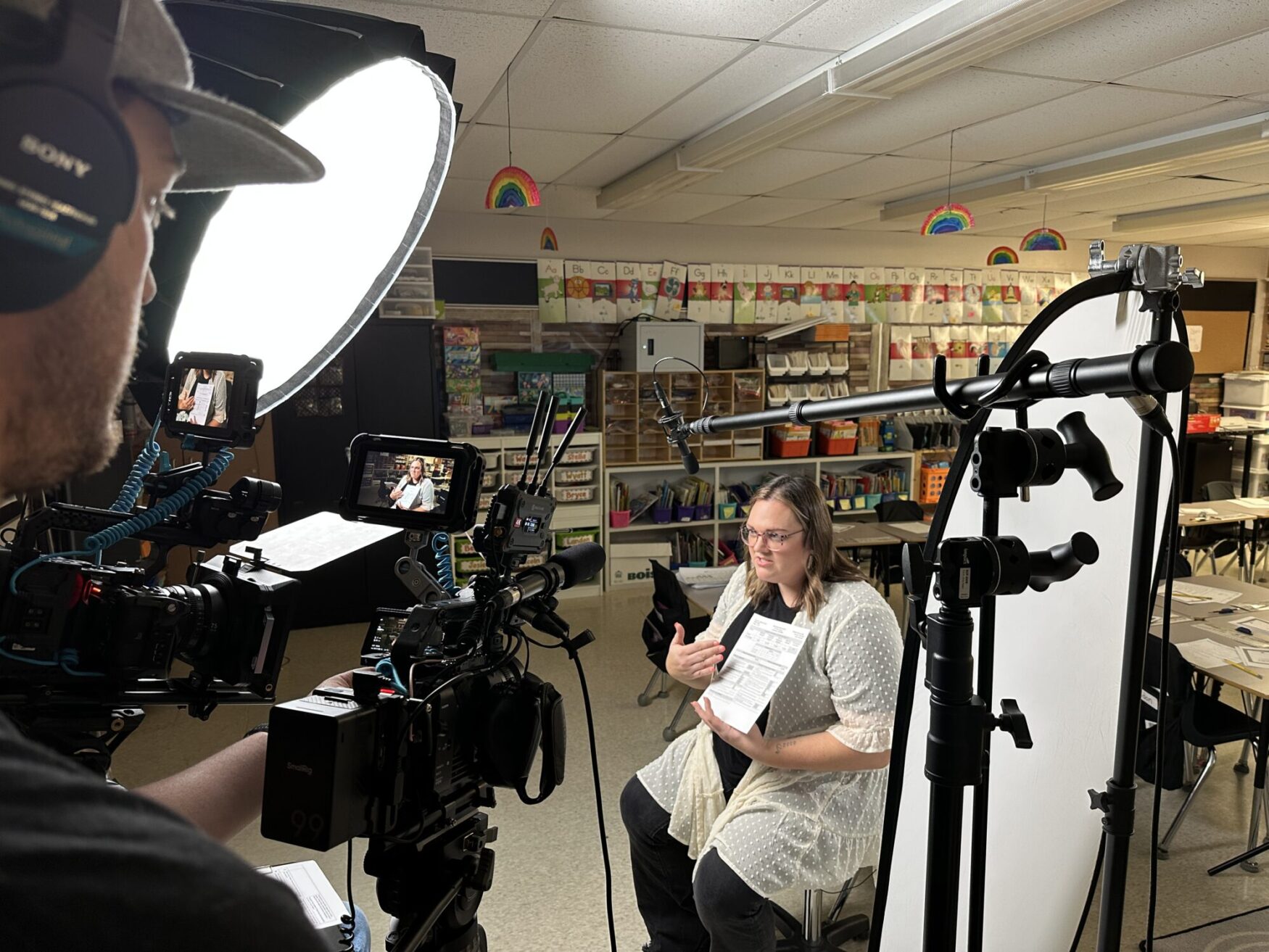 A behind-the-scenes look at a video interview setup in a colorful elementary school classroom. A woman sits on a stool, holding a document and speaking to the camera. A filmmaker, wearing headphones, operates a professional camera rig with an external monitor displaying the interview. Soft lighting and a boom microphone are positioned for optimal audio and video quality.