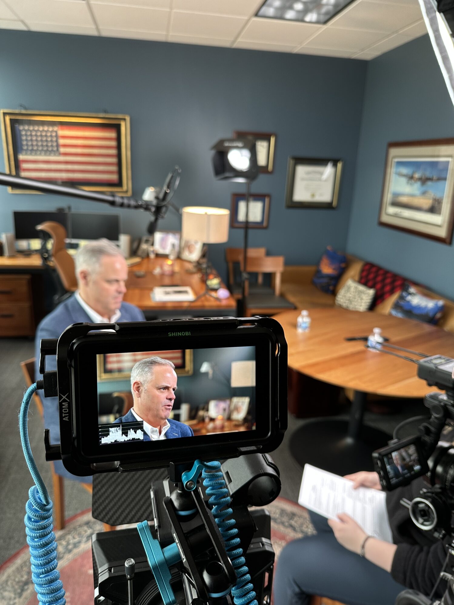 A professional interview setup in an office, with a man in a navy blazer speaking on camera. The image is framed through a camera monitor displaying the subject in focus, while the background includes an American flag, framed certificates, a wooden desk, and a cozy seating area with pillows. A boom microphone and studio lighting are visible, and the interviewer, holding a sheet of questions, is partially seen in the foreground.