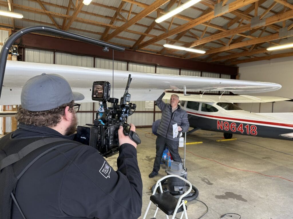 A cameraman wearing a Tellwell-branded jacket films a man inspecting or cleaning the wing of a small aircraft inside a hangar. The man, dressed in a gray jacket, holds a cloth and a checklist while standing on a stool. The aircraft, marked with the tail number "N96418," is parked under a wooden-beamed ceiling with bright overhead lights. The scene captures a behind-the-scenes look at an aviation-related video production.
