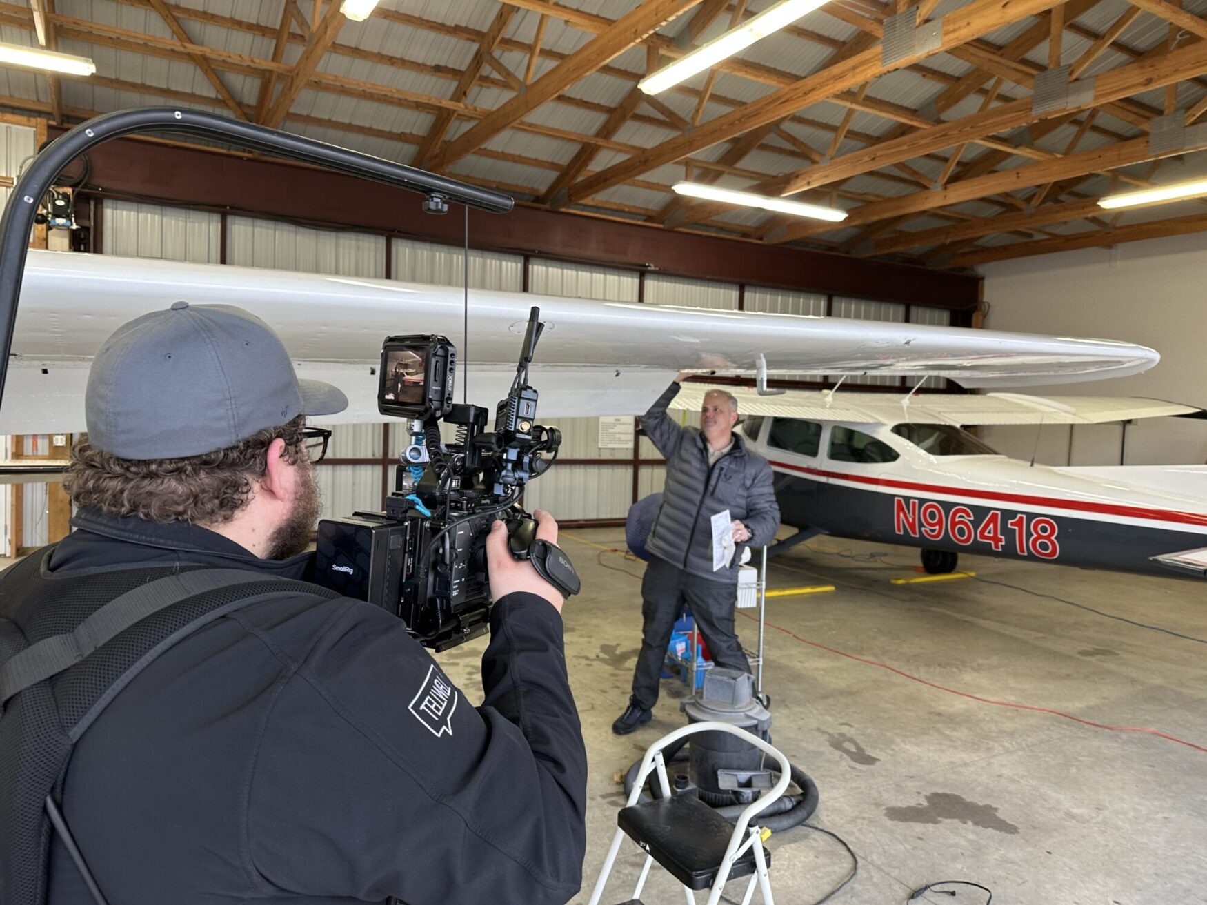 A cameraman wearing a Tellwell-branded jacket films a man inspecting or cleaning the wing of a small aircraft inside a hangar. The man, dressed in a gray jacket, holds a cloth and a checklist while standing on a stool. The aircraft, marked with the tail number "N96418," is parked under a wooden-beamed ceiling with bright overhead lights. The scene captures a behind-the-scenes look at an aviation-related video production.