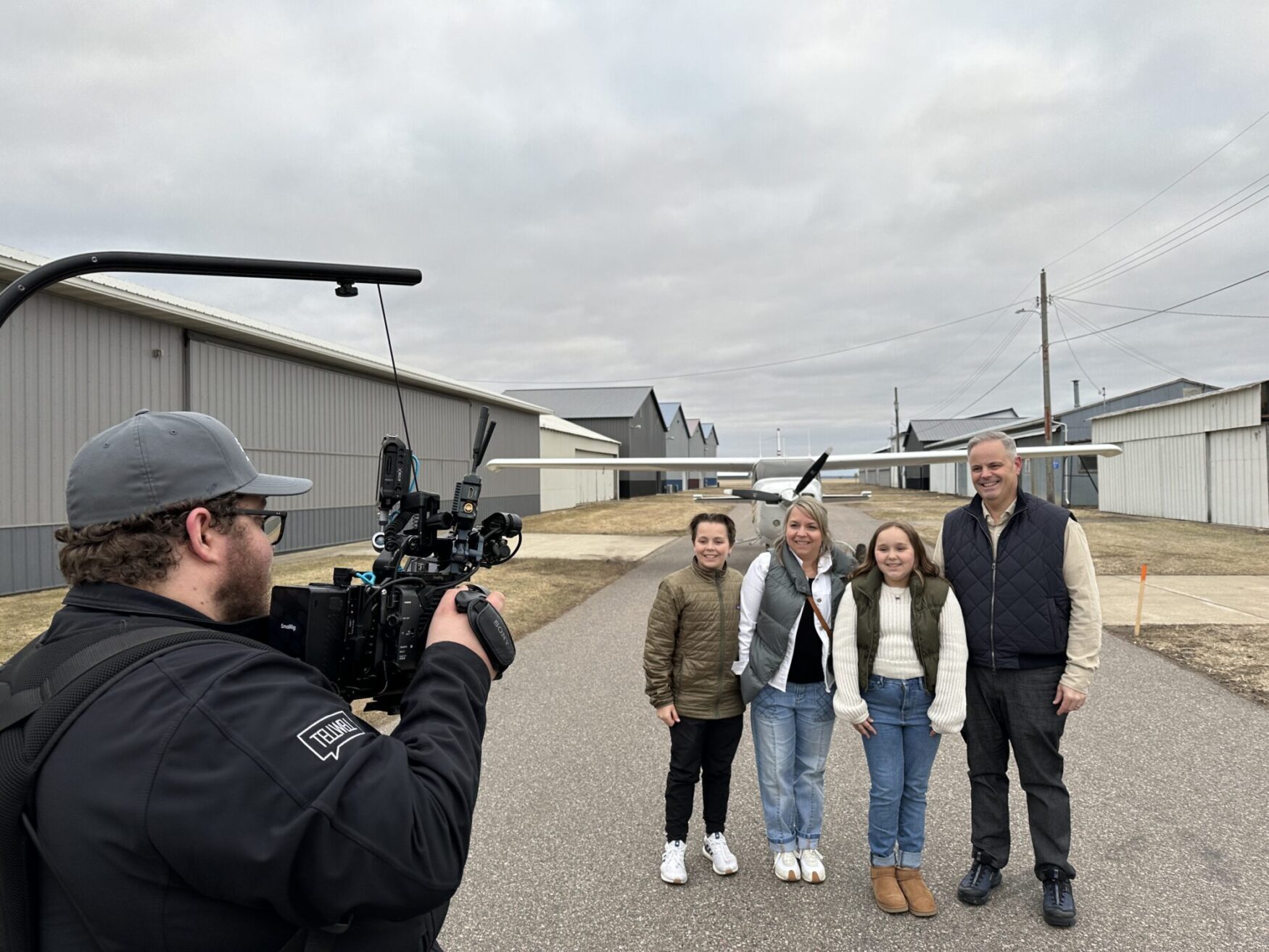 A film crew member operates a professional camera rig, capturing a family of four posing in front of a small airplane at an airfield. The family, dressed in warm clothing, smiles for the shot, standing between rows of metal hangars under an overcast sky. The cameraman wears a Tellwell-branded jacket and cap, focusing intently on the scene.