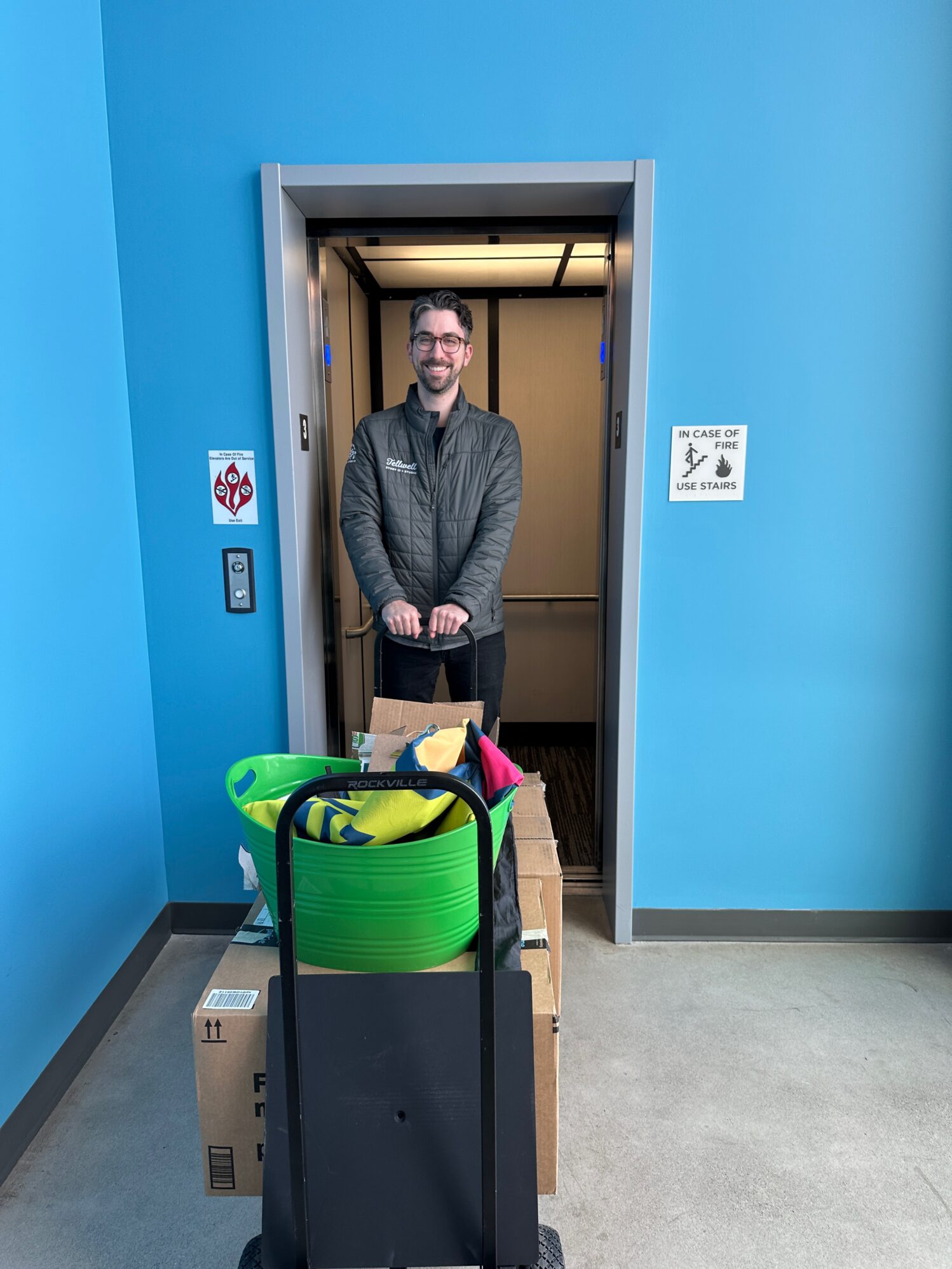 A man in a green jacket stands smiling inside an elevator with a cart full of supplies, including a bright green bin and cardboard boxes, in front of a vibrant blue wall.