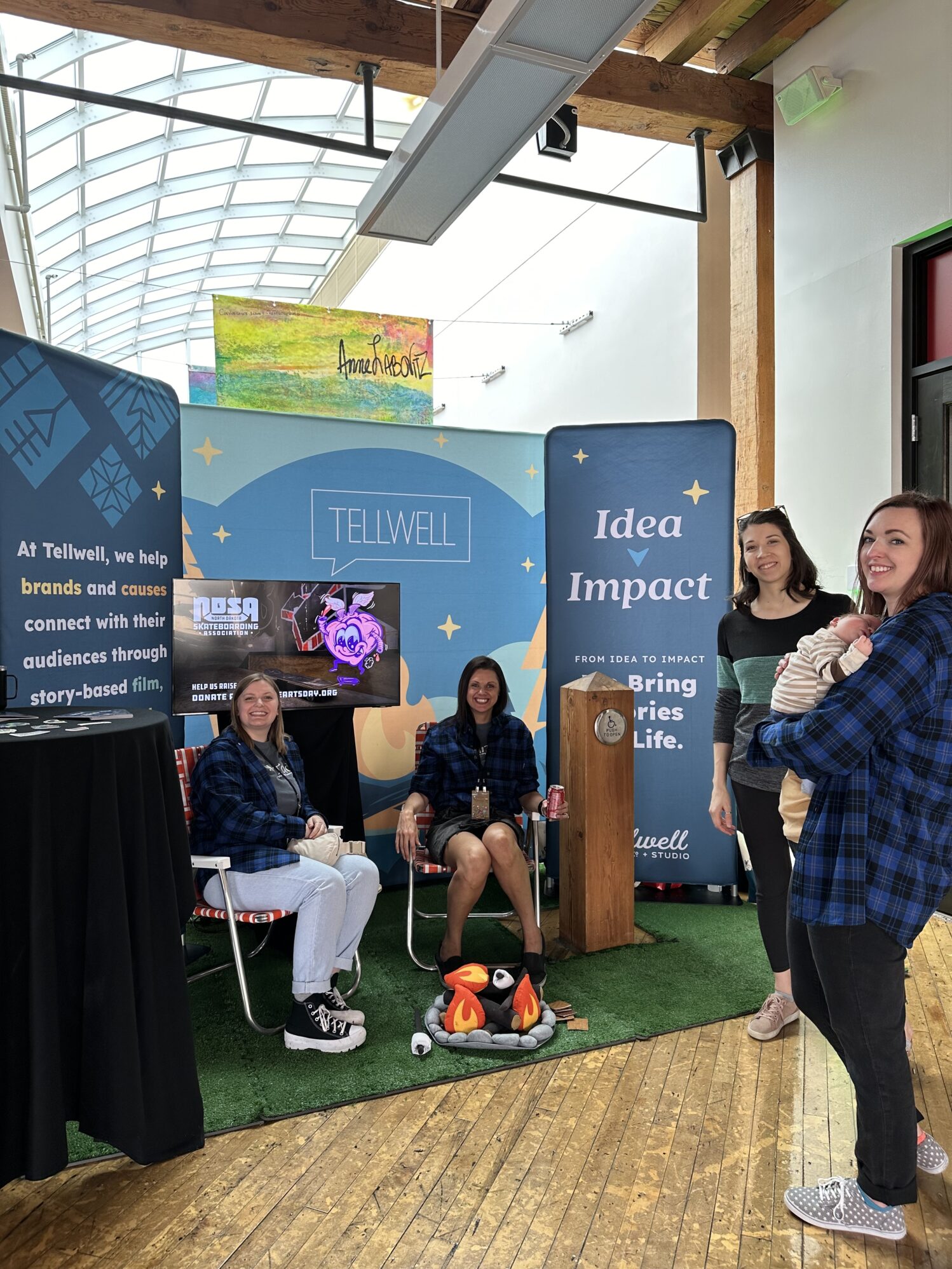 A group of women in blue flannels gather in front of a colorful Tellwell event booth with a faux campfire setup, branded backdrops, and a sign reading 'Idea to Impact'.