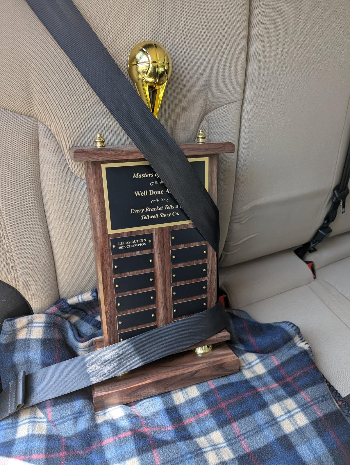 A large wooden trophy with a golden basketball topper, labeled 'Well Done Award', is safely buckled in with a seatbelt in the back seat of a car, resting on a blue plaid blanket.