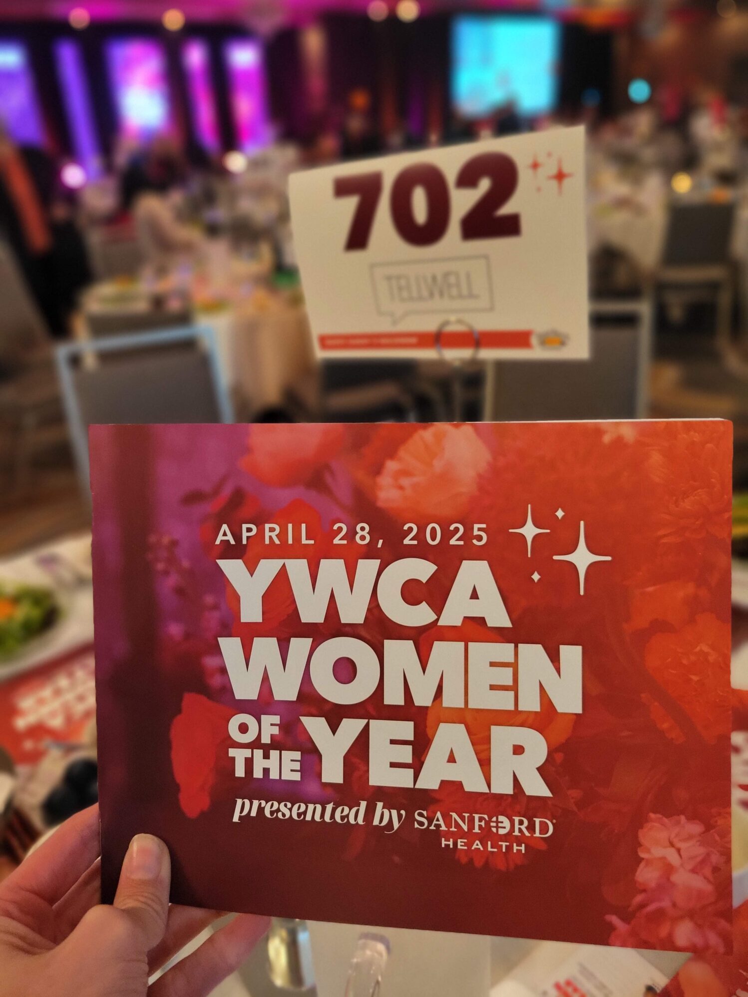 A person holds a vibrant event program that reads "April 28, 2025 – YWCA Women of the Year presented by Sanford Health" in bold white text against a red and floral background. In the blurred background, round banquet tables are set for dinner in a warmly lit event space. A table number sign reading “702” with the Tellwell logo is prominently visible.