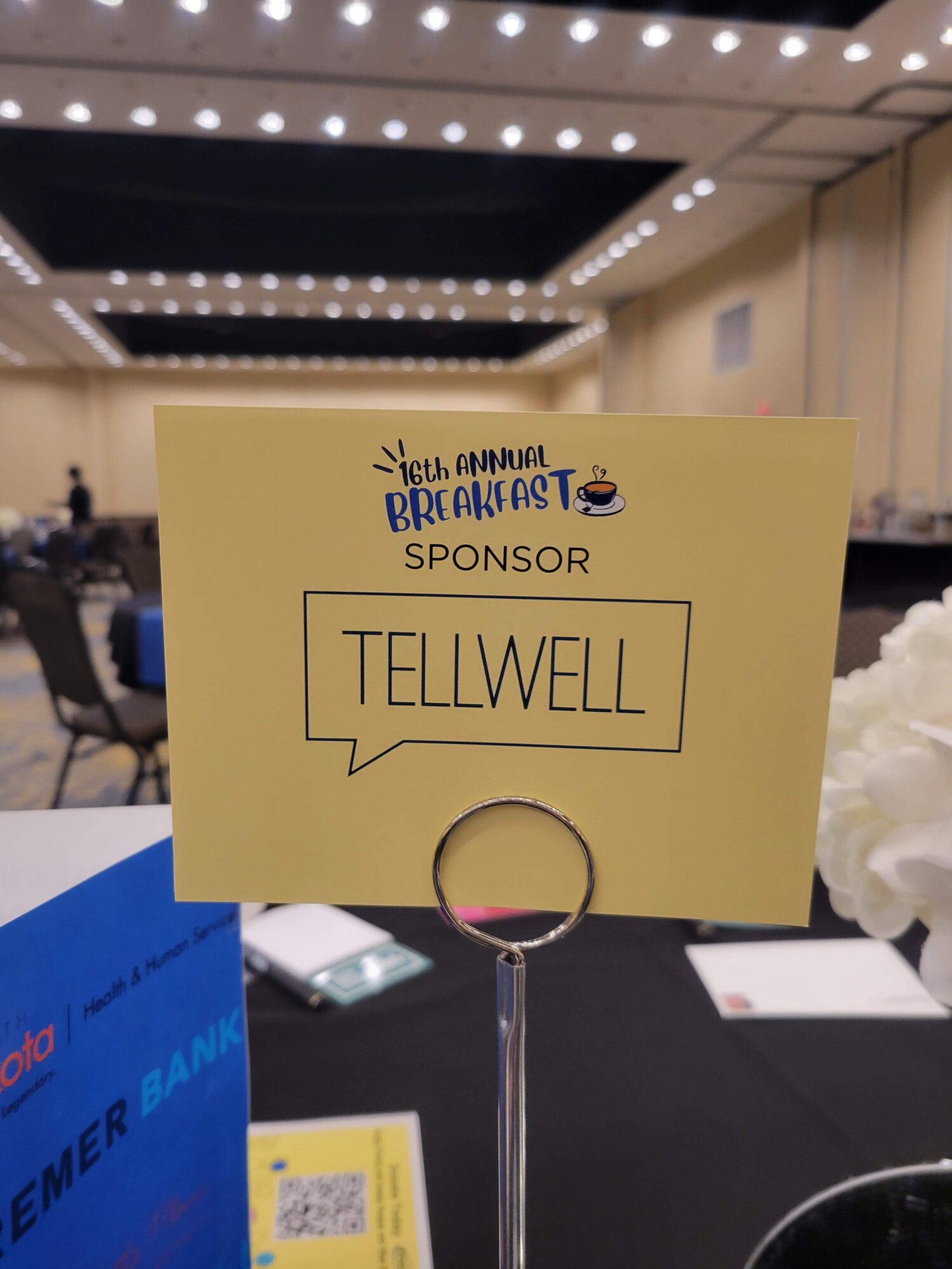 A yellow sponsor card on a metal holder reads “16th Annual Breakfast – SPONSOR – TELLWELL” with a small coffee cup graphic. The card is placed on a black tablecloth in a large, well-lit conference room with high ceilings and rows of ceiling lights. Other event materials and flowers are partially visible on the table.