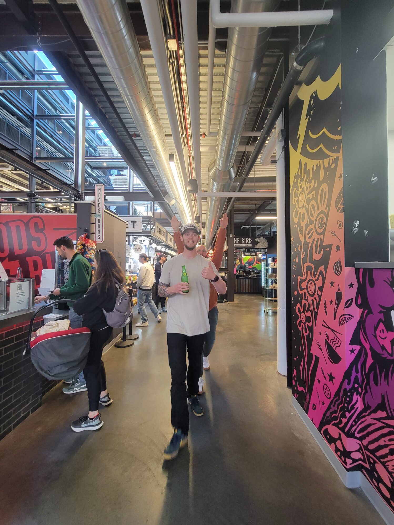 A man giving a thumbs up holding a green, glass bottle. He is in a cafeteria-like space which is industrial and colorful.