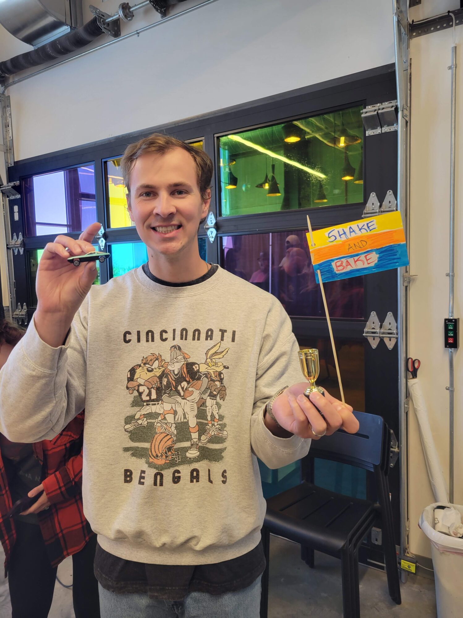 A man holds a small toy car, a small trophy, and a yellow, orange and blue flag that reads “Shake and bake”. He is smiling as he just won the award.
