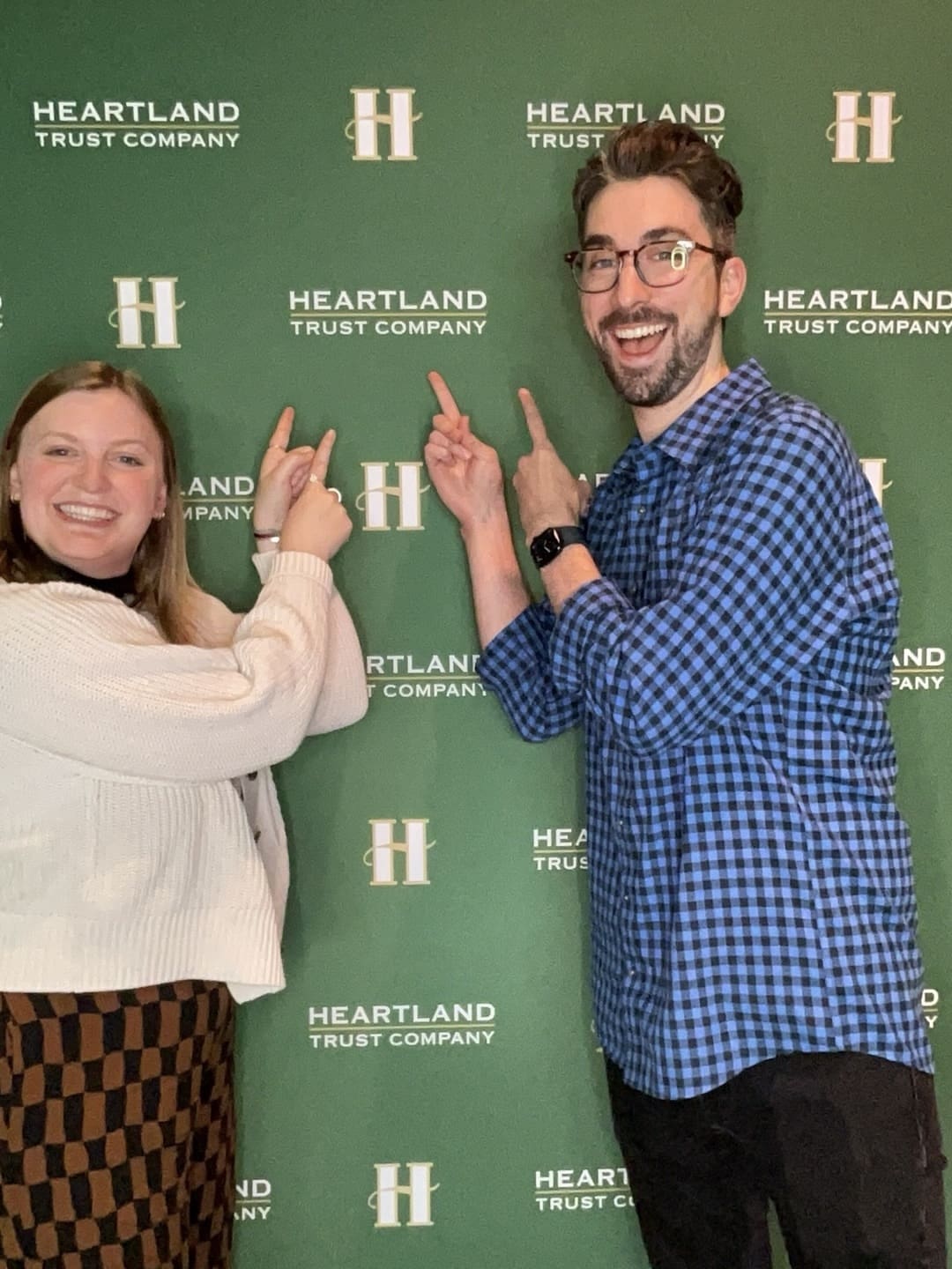 A man and a woman stand in front of a photo backdrop. The backdrop is dark green and reads “Heartland Trust Company”. They are both smiling and pointing at the test, showing their excitement to be at the event for this company.