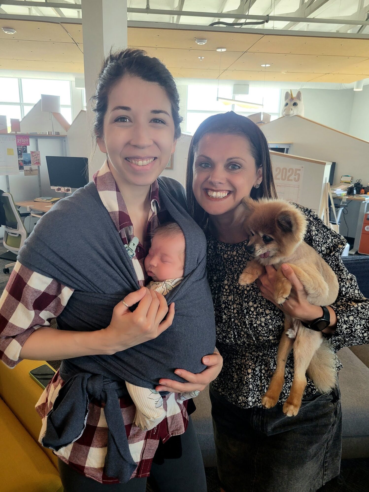 Two smiling women standing indoors in a modern office space. The woman on the left is wearing a plaid shirt and has a newborn baby wrapped snugly against her chest in a dark gray baby carrier. The woman on the right, wearing a patterned blouse, is holding a small fluffy dog. In the background are desks, chairs, and a calendar marked '2025.' A large stuffed animal head resembling a dog peeks over the top of a structure behind them.