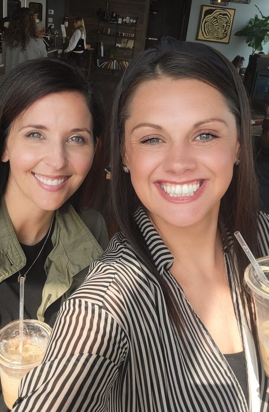 Two women smile brightly while taking a selfie inside a modern coffee shop. The woman on the left has straight dark hair and wears a green jacket over a black top. The woman on the right has long dark hair, a headband, and a black-and-white striped blouse. Both are holding iced coffee drinks. In the background, people can be seen near the counter, and the space features industrial ceilings, pendant lighting, and modern decor.