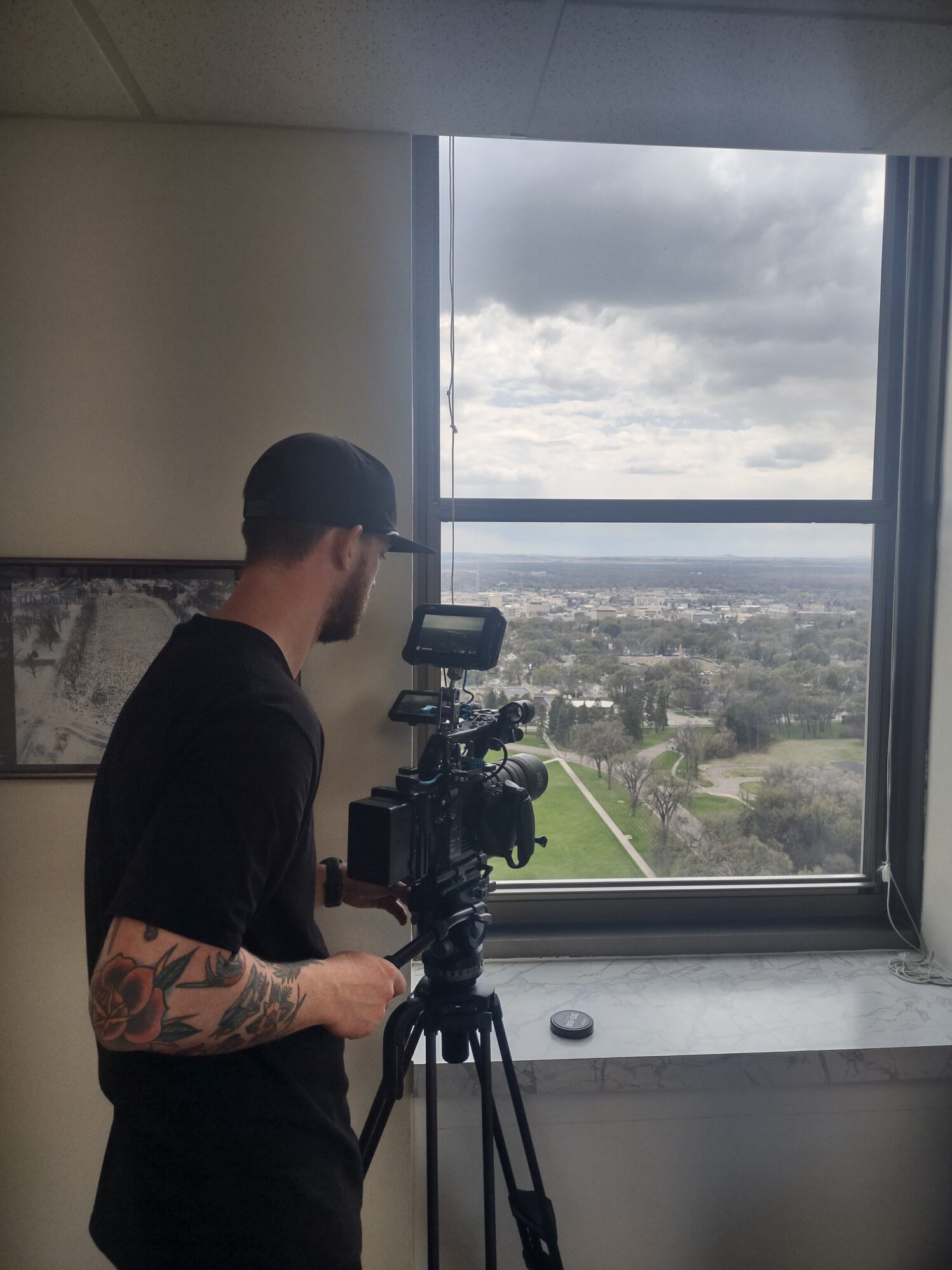 A man with tattooed arms and a backward baseball cap operates a professional video camera on a tripod inside a high-rise building. He is filming out a large window that overlooks a cityscape and green parkland under a cloudy sky.