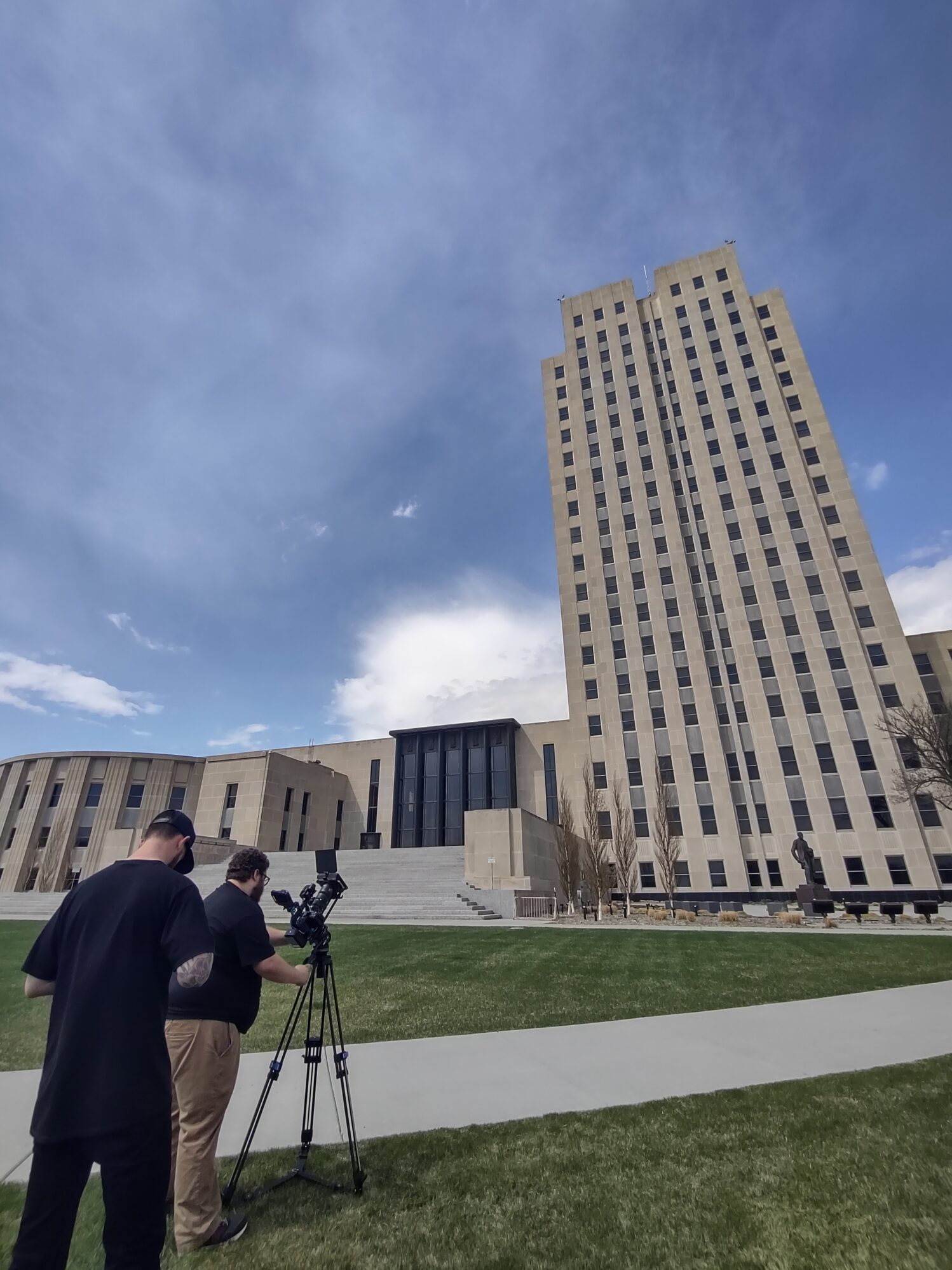 Two people operate a professional video camera on a tripod outside the North Dakota State Capitol building in Bismarck. The tall Art Deco tower rises into a blue, lightly clouded sky, while the expansive lawn and wide steps leading to the entrance create a dramatic foreground.
