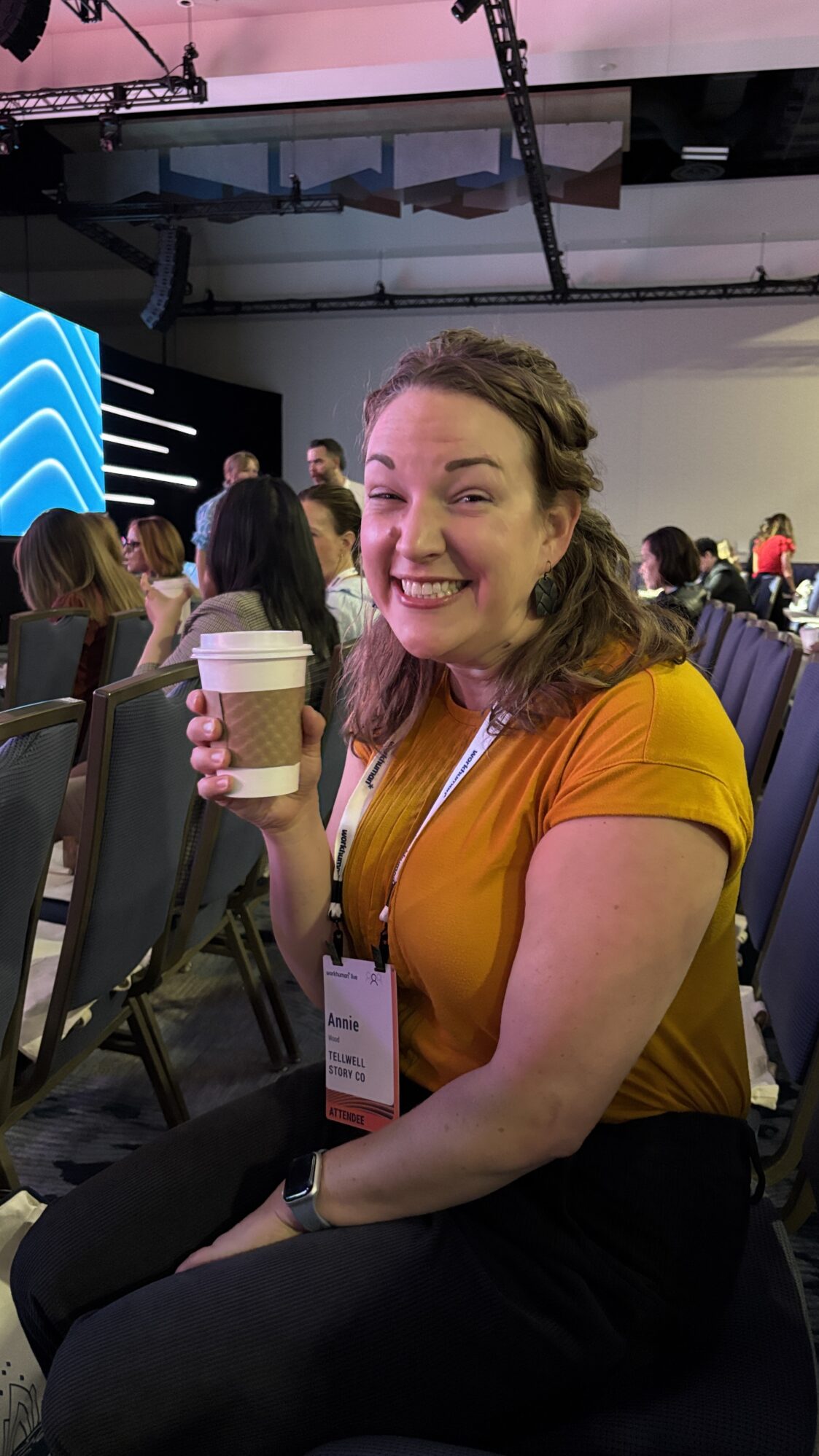 A woman wearing a yellow t-shirt smiles at the camera. She is holding a cup of coffee and wearing a lanyard with a name badge, suggesting she is attending a conference. There are several rows of chairs surrounding her. Her expression shows excitement and happiness.