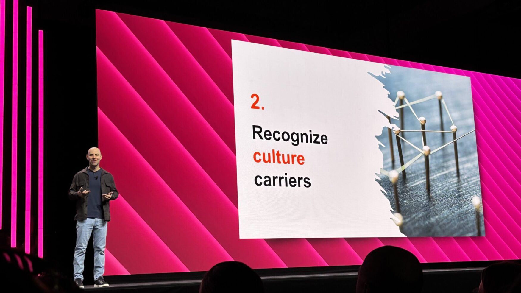 A speaker stands on stage in front of a large screen with a bold pink and black backdrop. The slide behind him reads “2. Recognize culture carriers” with the word “culture” highlighted in red. The slide also includes an abstract image of gold pins connected by string, symbolizing a network. The speaker is mid-presentation, gesturing with both hands.