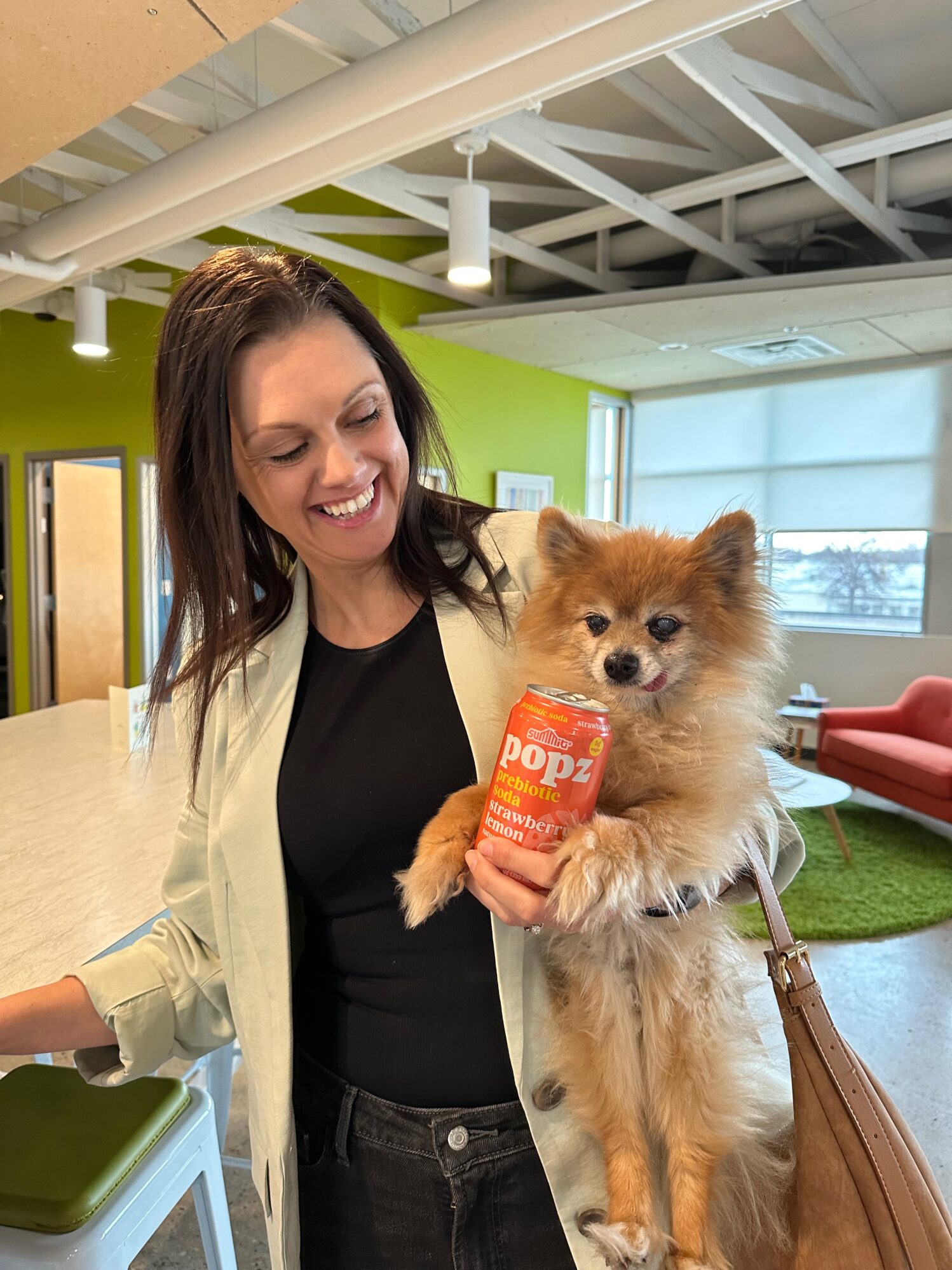 A woman with long brown hair, wearing a light jacket over a black top and dark jeans, smiles while holding a fluffy Pomeranian dog. The dog is perched in her arm and is holding a can of "Popz prebiotic soda" in strawberry lemon flavor. They are indoors in a brightly lit modern office space with green and orange decor.