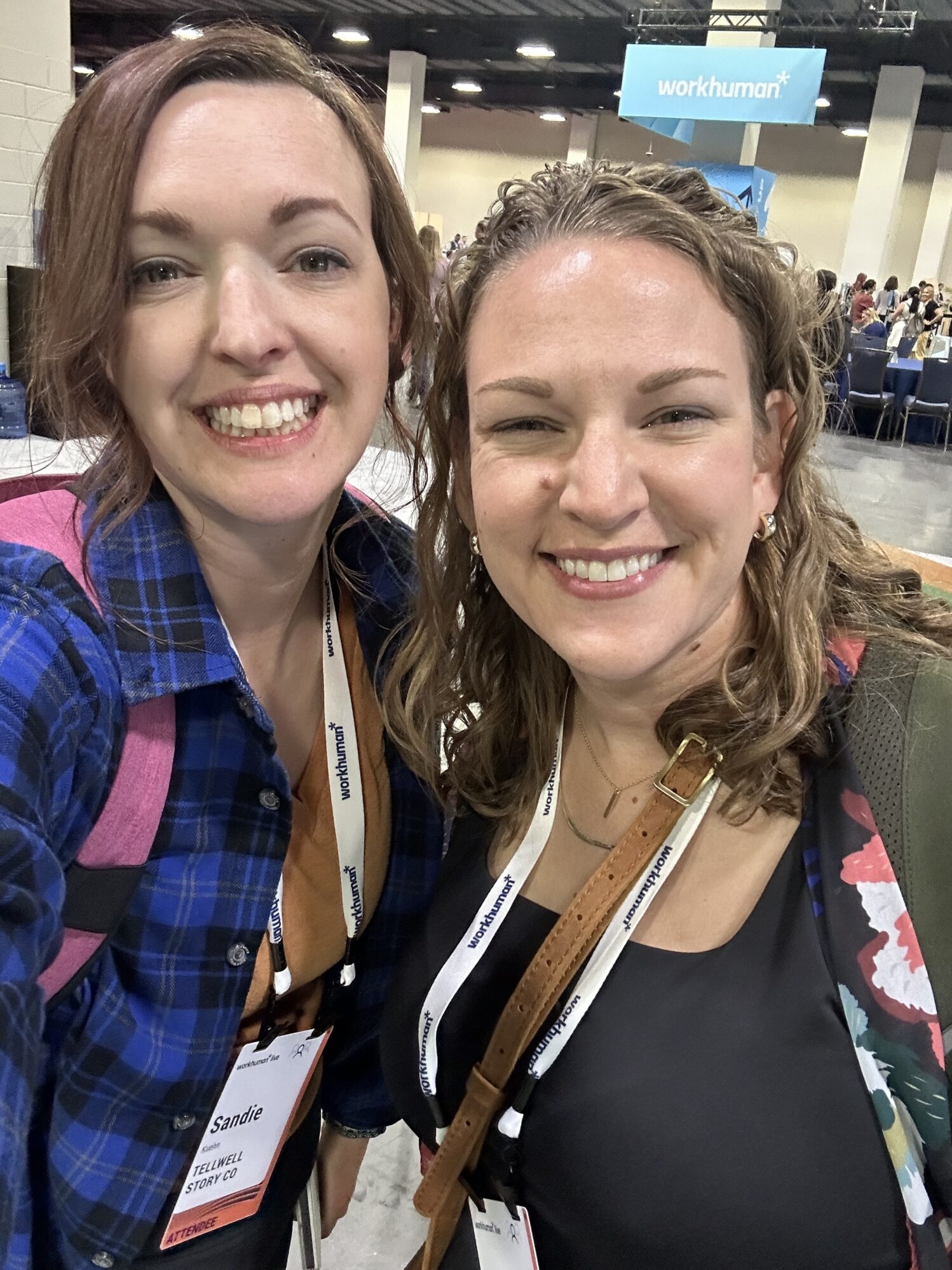 Two women smile for a selfie at an indoor conference. They are both wearing name badges and lanyards that say “workhuman.” One wears a blue plaid shirt and pink backpack, and the other wears a black top with a colorful bag strap. A “workhuman” sign hangs in the background above tables filled with people.