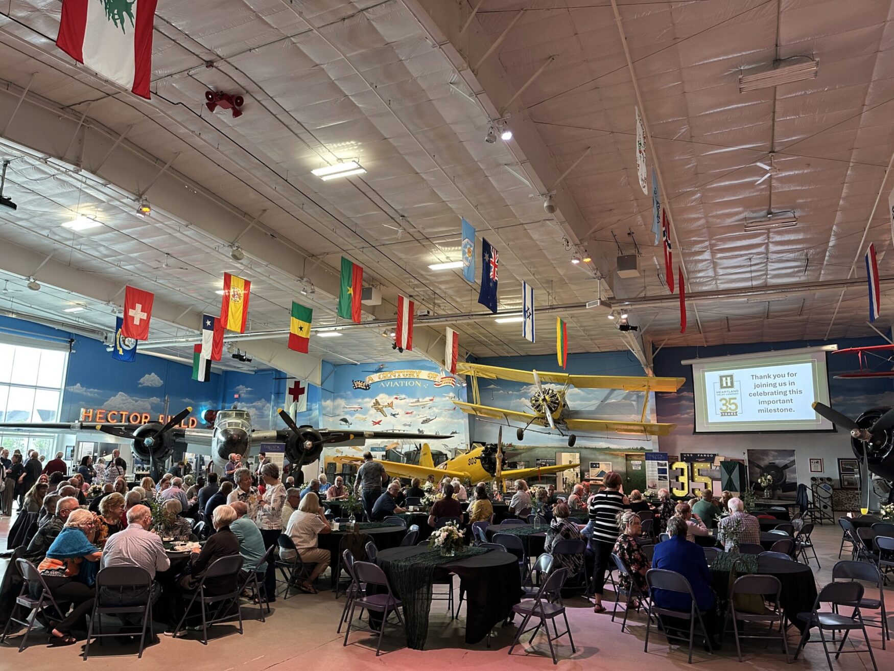 An event with several tables filled with attendees takes place inside an air museum. There are flags of different countries hanging from the ceiling and planes spread throughout the space. There is a screen that reads “Heartland Trust Company. Thank you for joining us in celebrating this important milestone.”