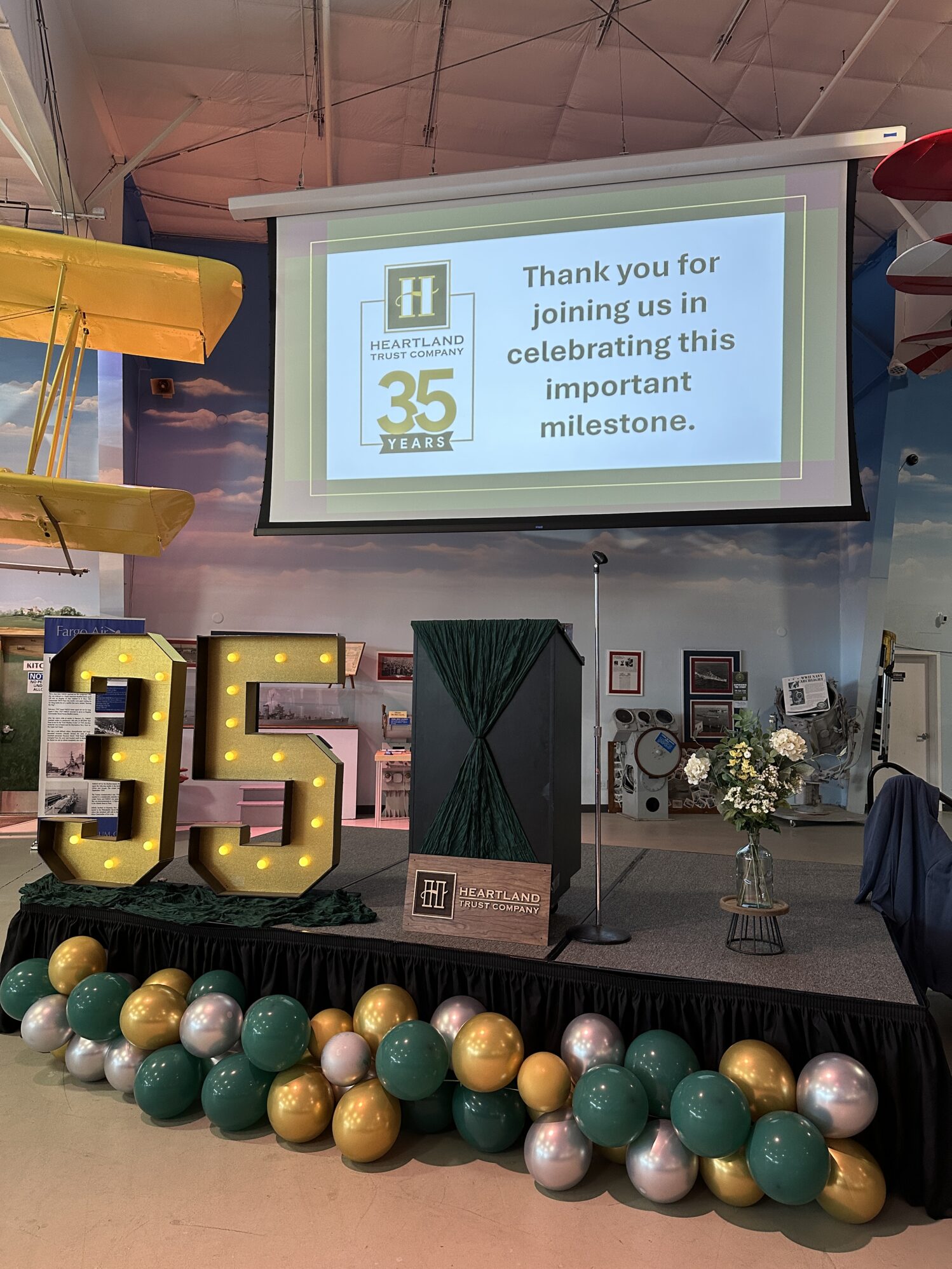 A decorated stage setup for the Heartland Trust Company’s 35th anniversary celebration. A large screen displays the message: 'Thank you for joining us in celebrating this important milestone,' along with the Heartland Trust Company logo and '35 Years.' The stage features a podium wrapped in green string with the company’s logo, a large illuminated '35' sign, and a bouquet of white flowers in a glass vase. Green, gold, and silver balloons line the front of the stage. The event is set in an aviation-themed venue, with a yellow biplane and airplane décor visible in the background.