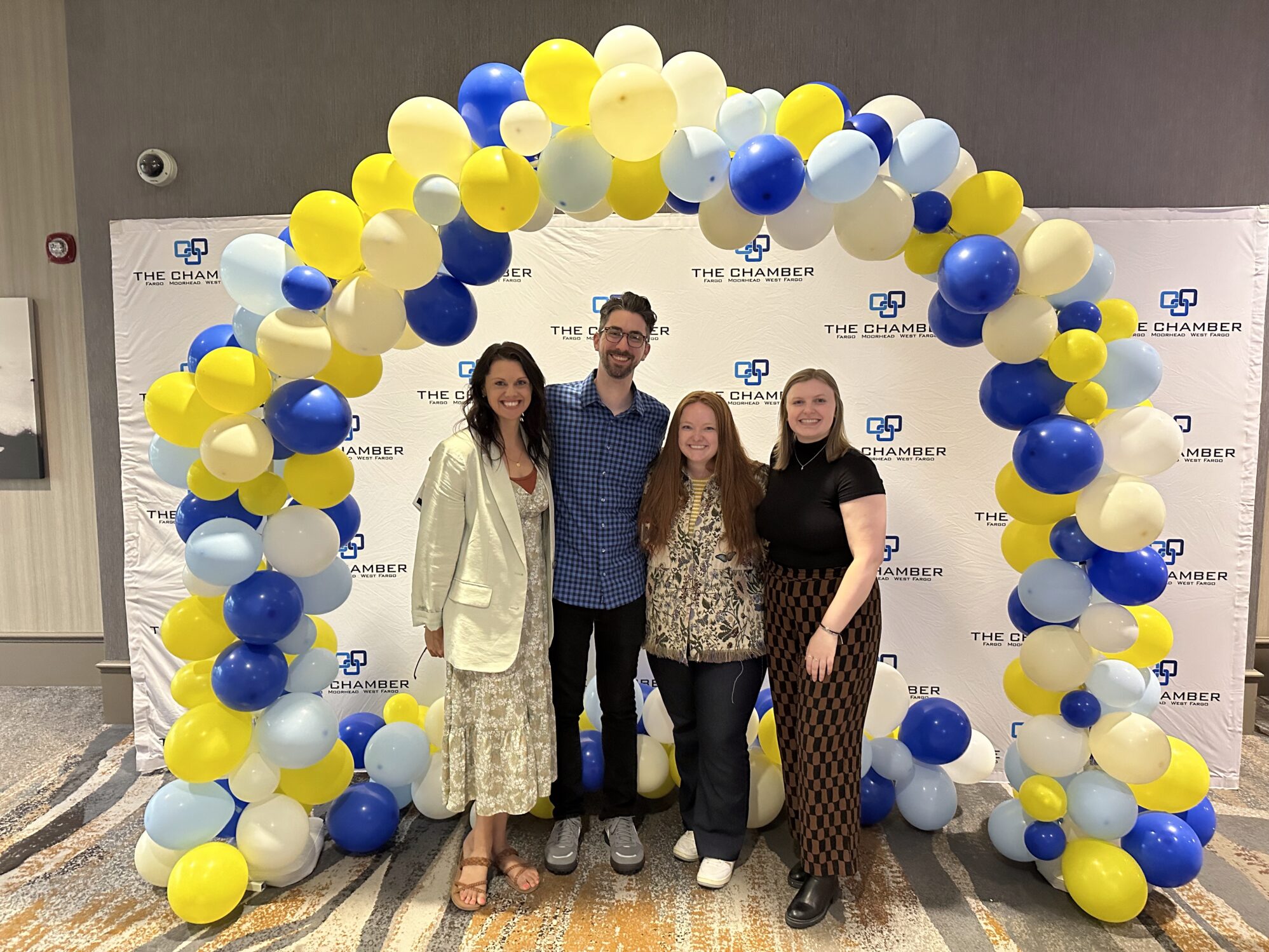 Three women and one man stand for a photo under a balloon arch created with yellow, white, and blue balloons. They appear to be at an event. There is a backdrop that reads “The Chamber Fargo Moorhead West Fargo”.