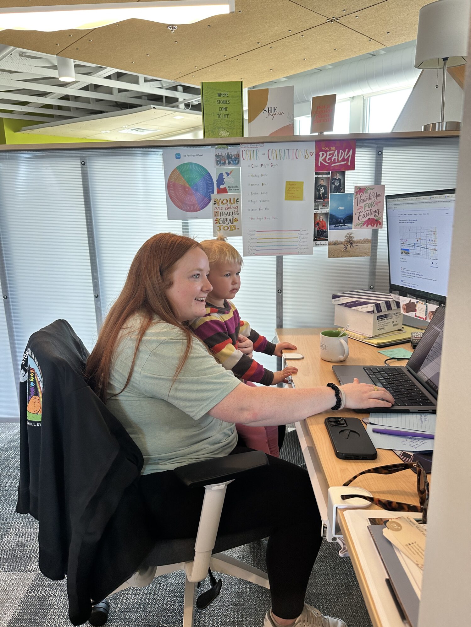 A woman with red hair is sitting at a desk in an office chair. A small blonde child is sitting on her lap as she shows her something on the computer screen.