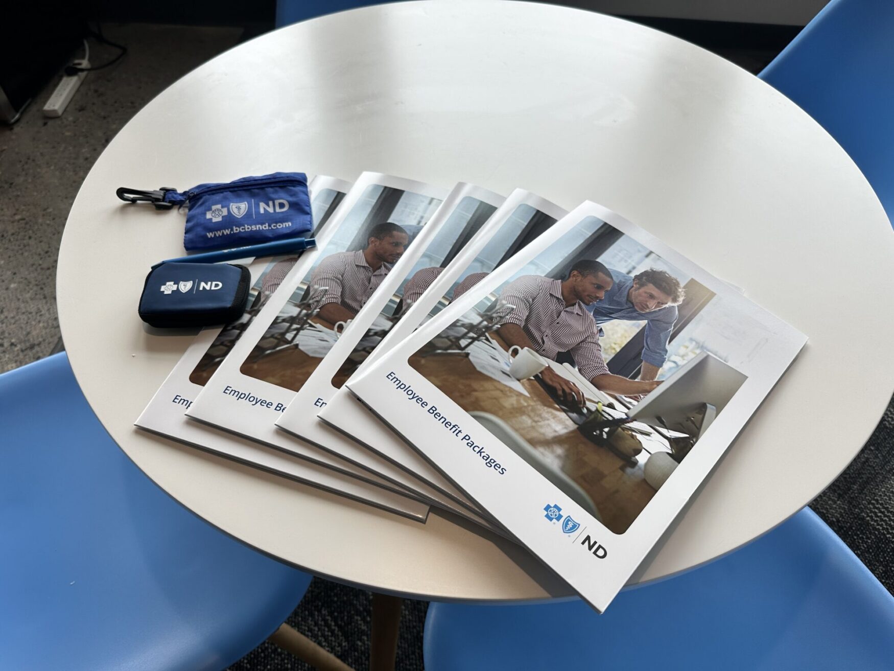 Several folders labeled "Employee Benefit Packages" sit in a circular white table. Next to the folders are two small blue bags.