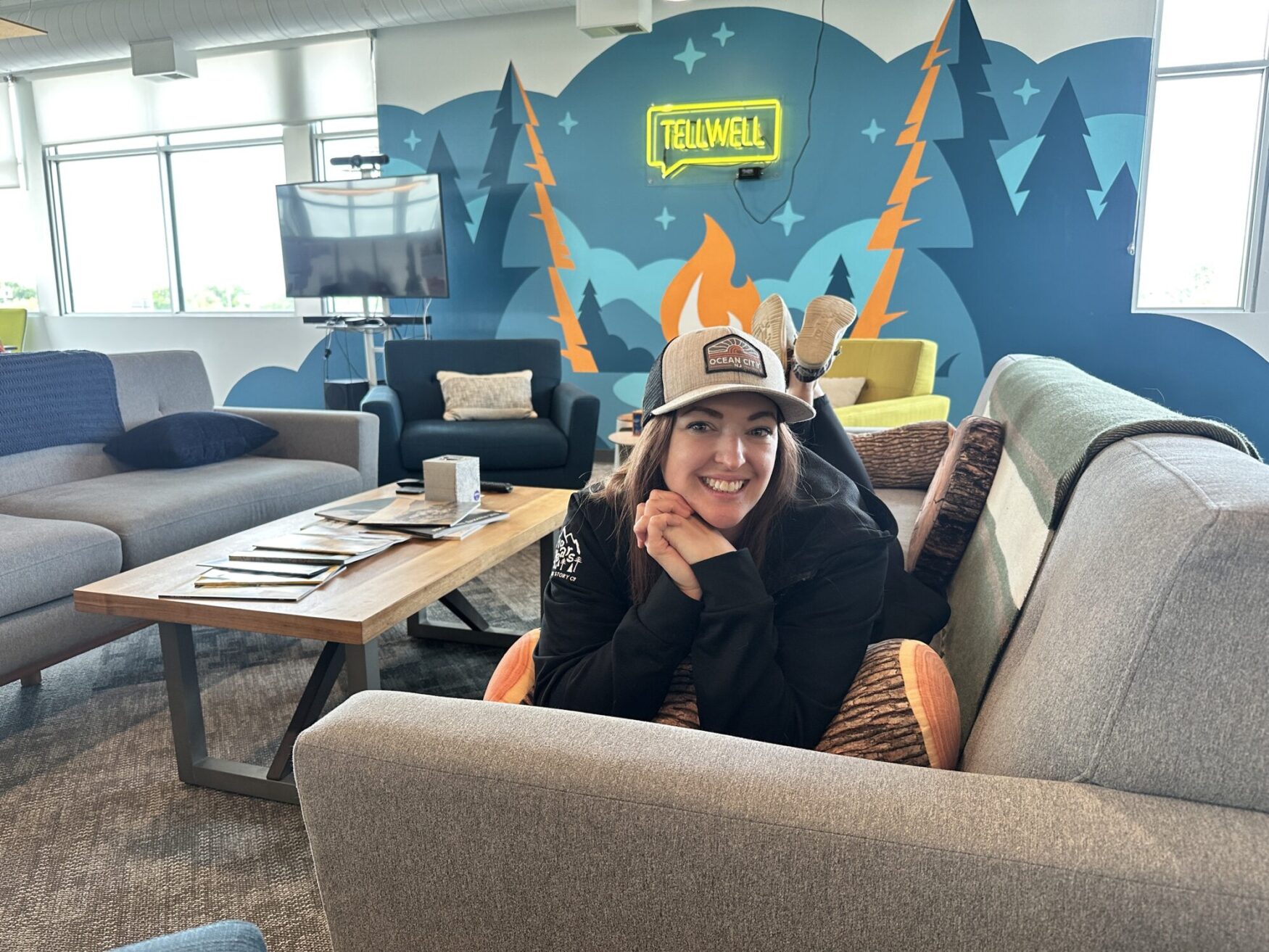 A woman smiles while lying on a couch with her chin resting on her hands and feet playfully kicked up behind her. She is wearing a gray "Ocean City" baseball cap and a black jacket. Behind her is a cozy office lounge with modern couches, a wooden coffee table with magazines, and a mural of pine trees, mountains, and a campfire. A bright yellow neon "TELLWELL" sign glows above the mural.