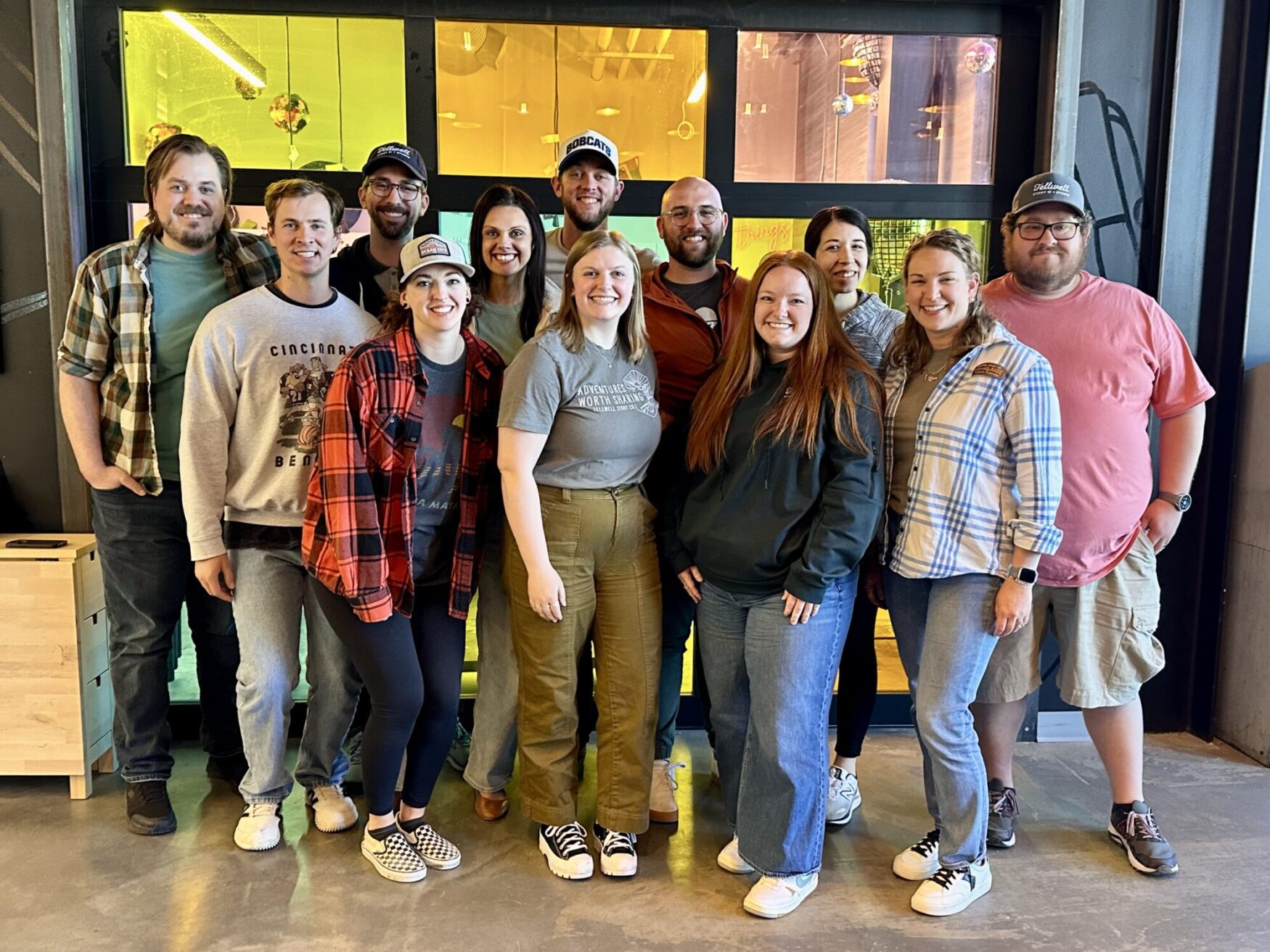 A group of 12 stand in front of multi-colored glass windows. There is a mix of men and women, they are all smiling.